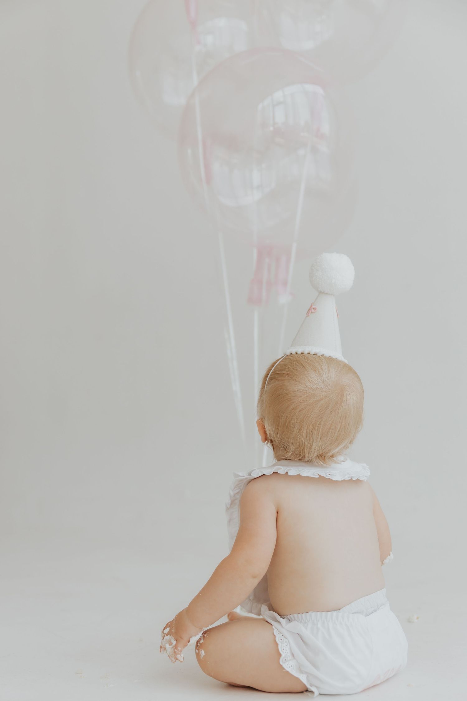Baby wearing white diaper sits with back turned against white background.