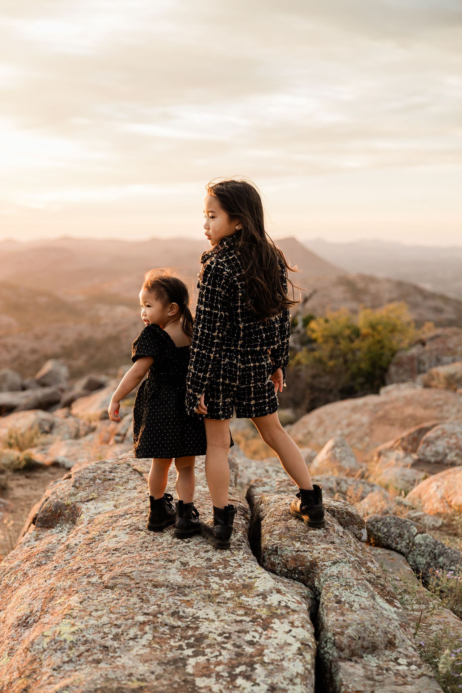 Two figures sitting on rocky cliffs overlooking mountains at golden hour.