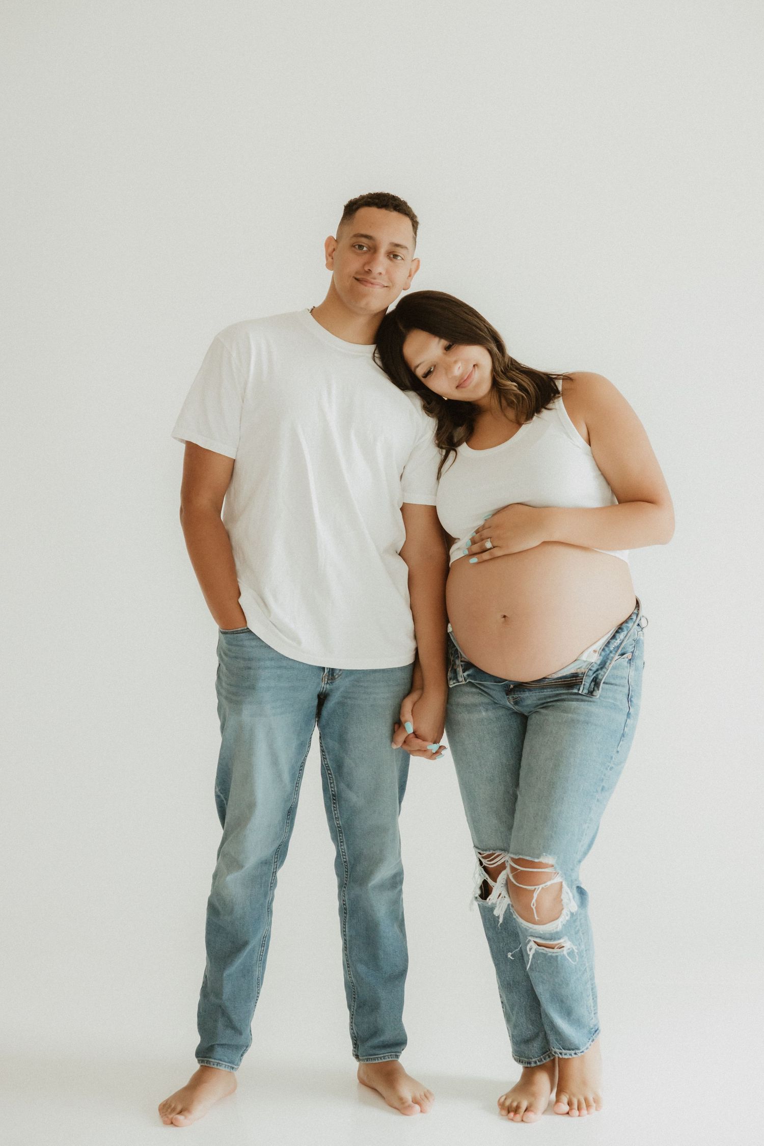 Minimalist maternity photos of a couple in jeans and white shirts against a white background.