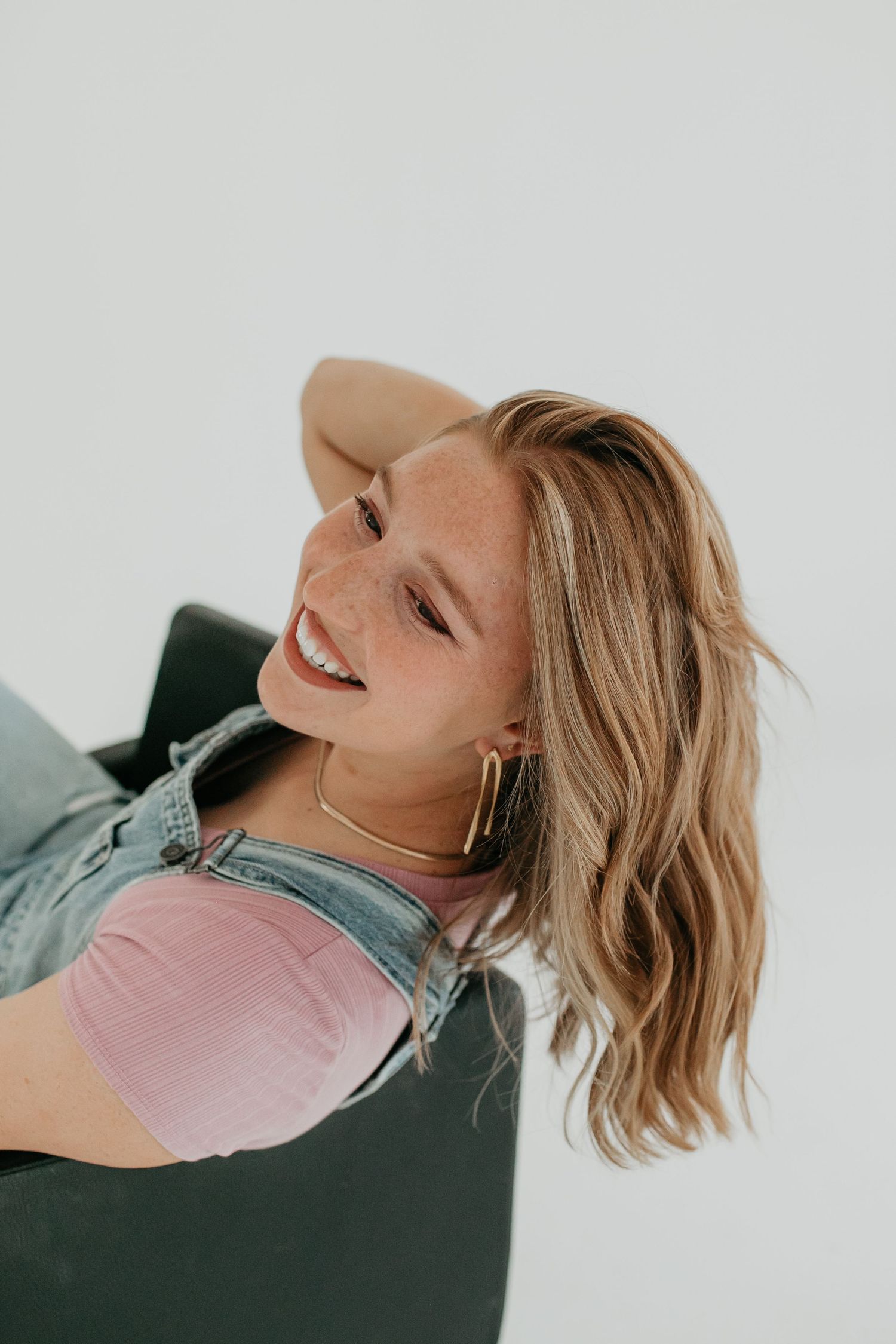 A person in a pink top reclines and laughs while laying on a dark chair against a light background.