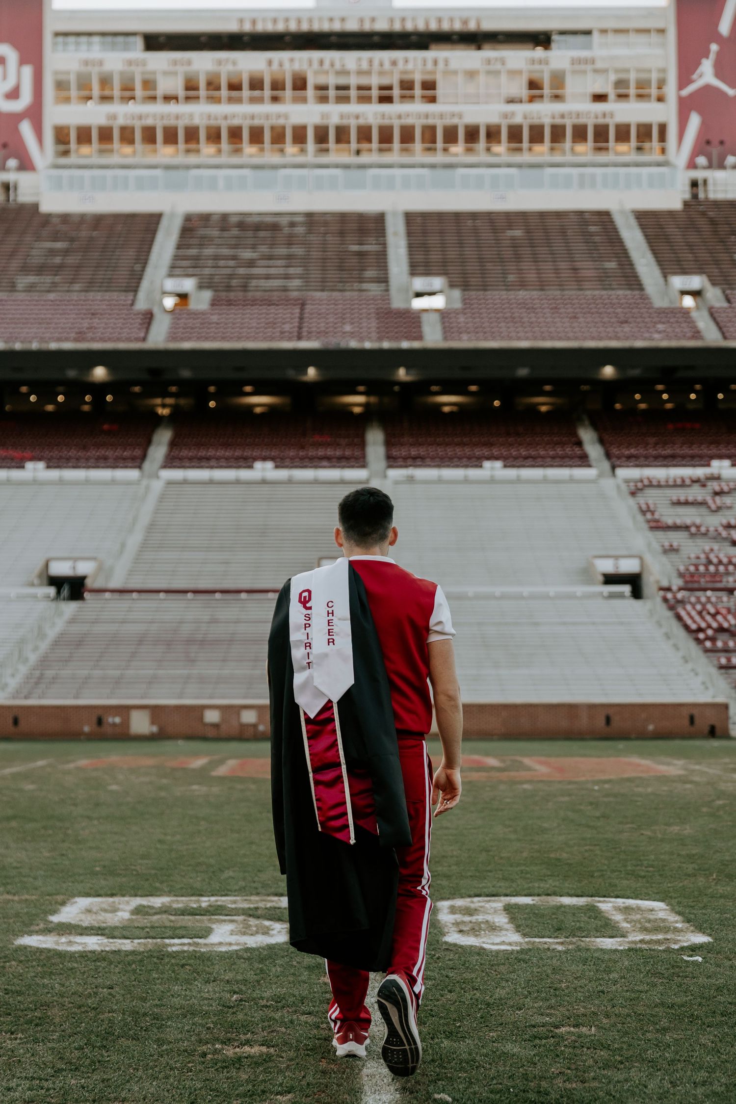 Graduate in graduation attire walks across football field at sunset.