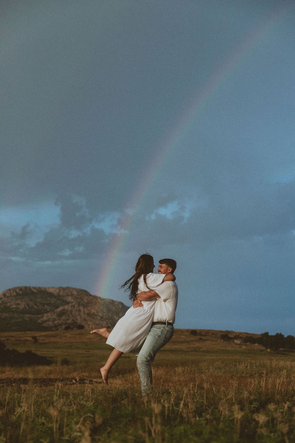 Couple dancing in field with dramatic rainbow and stormy sky backdrop.