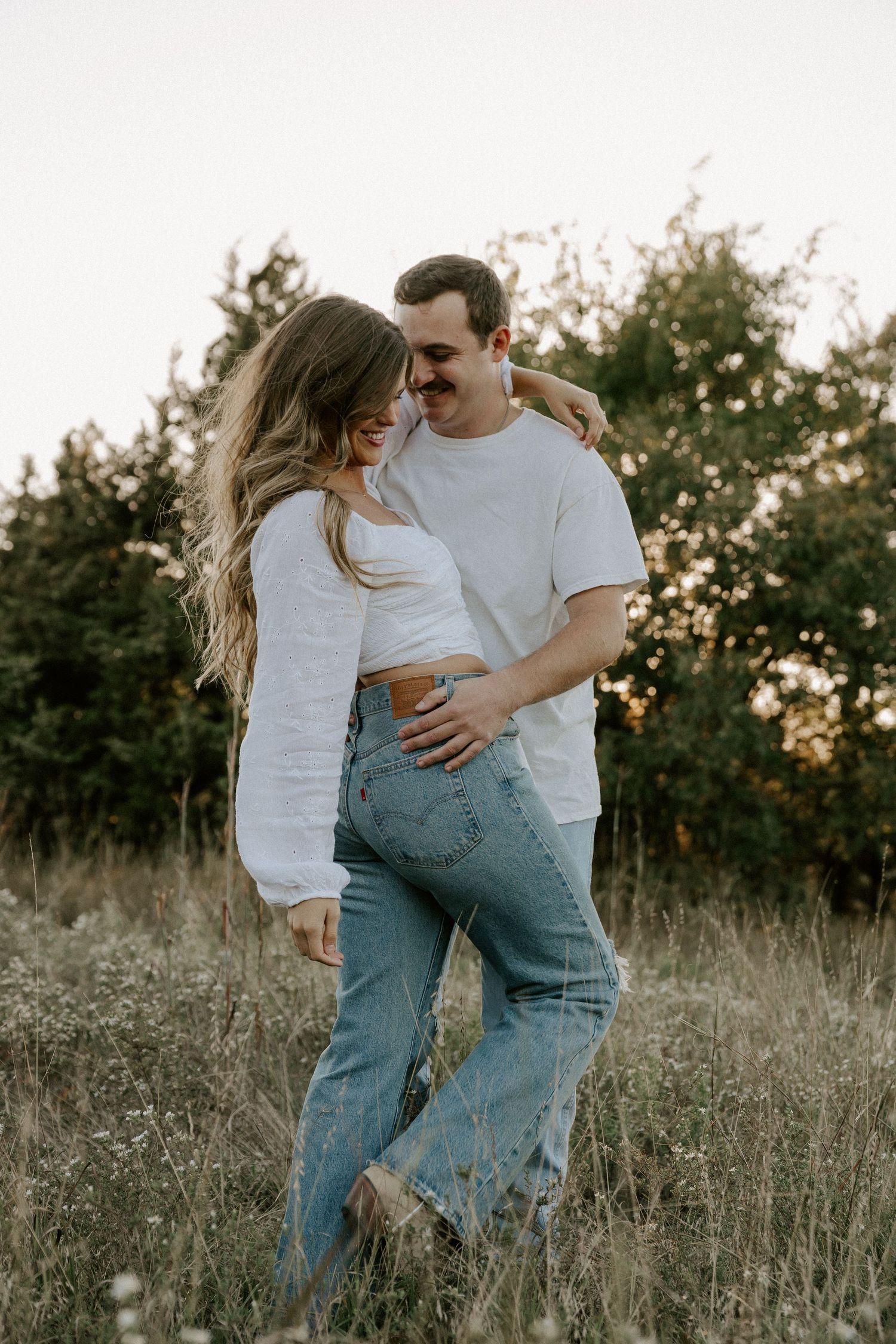 Casual couple in white shirts and jeans sharing romantic moment in sunset field.