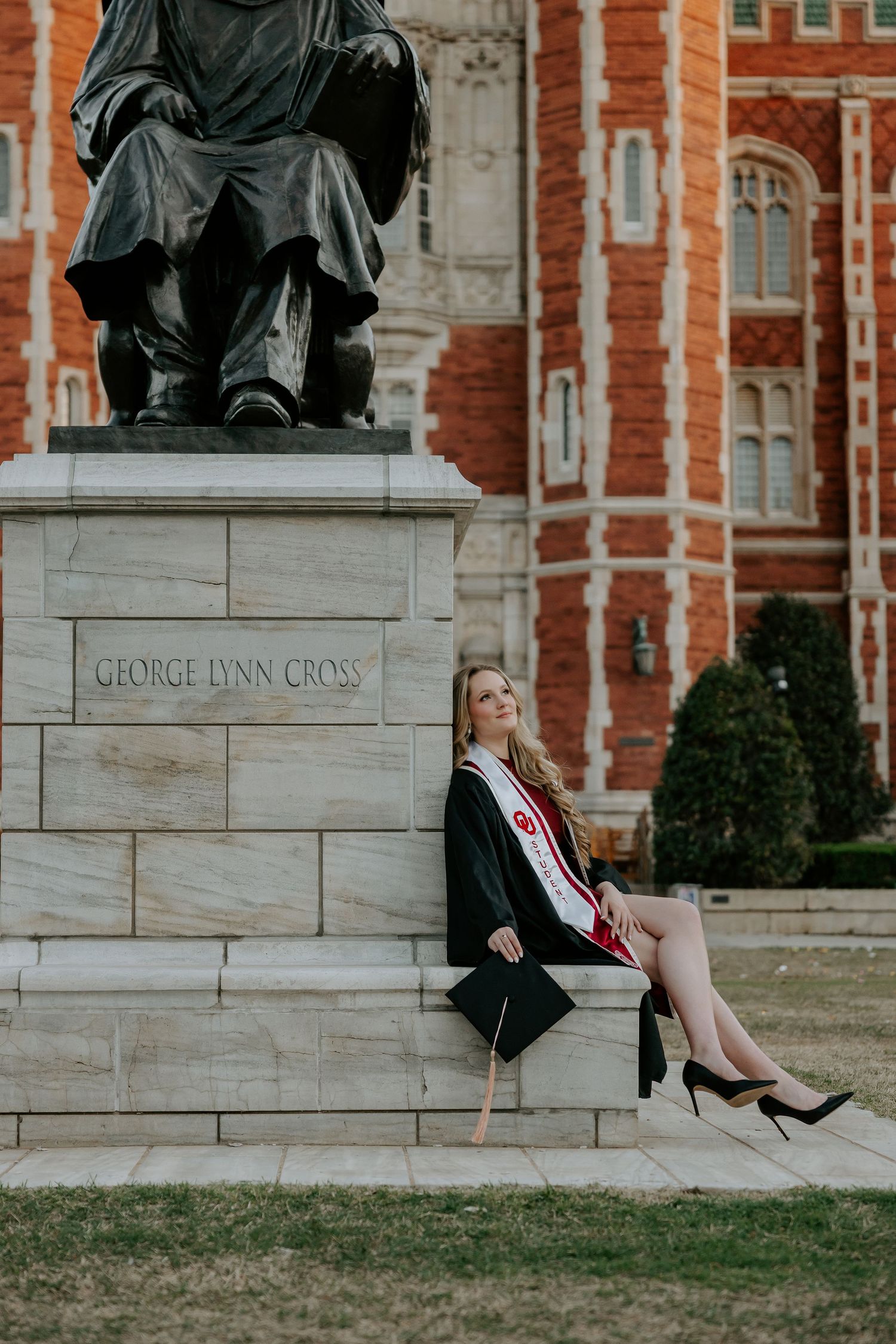 Graduate in formal attire sitting on stone monument near brick building.