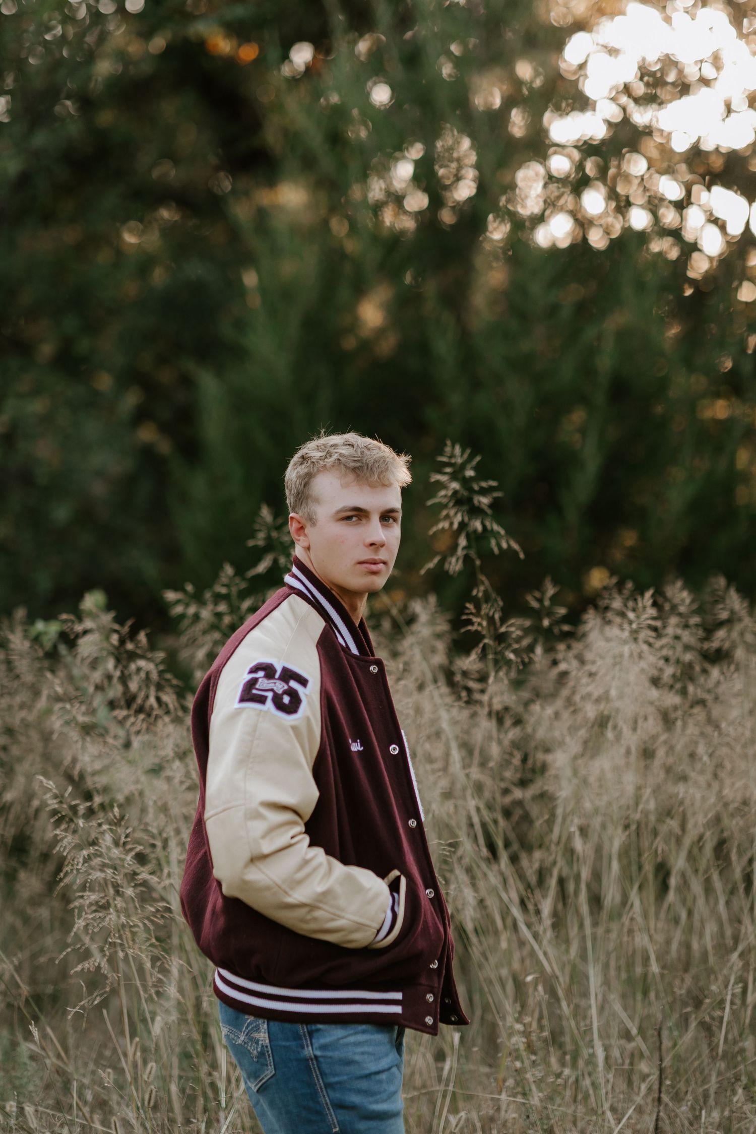 Student wearing varsity letterman jacket poses in tall grass during golden hour.
