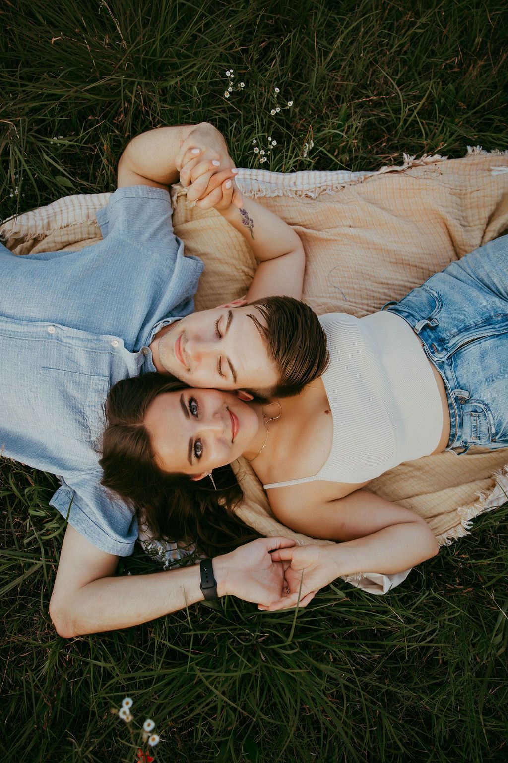 Person lying on blanket in grass with flowers, wearing denim and white clothing.