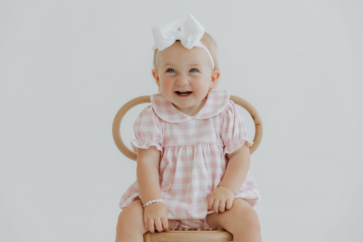 Baby in pink gingham dress sitting against white background.