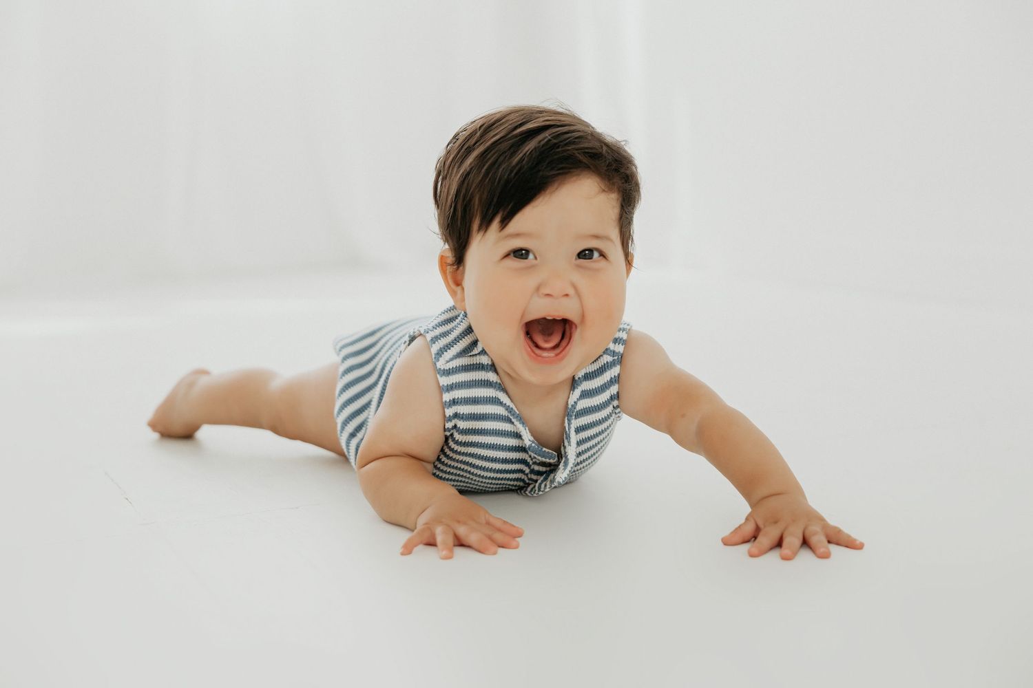 Baby in striped outfit crawls and smiles on white floor.