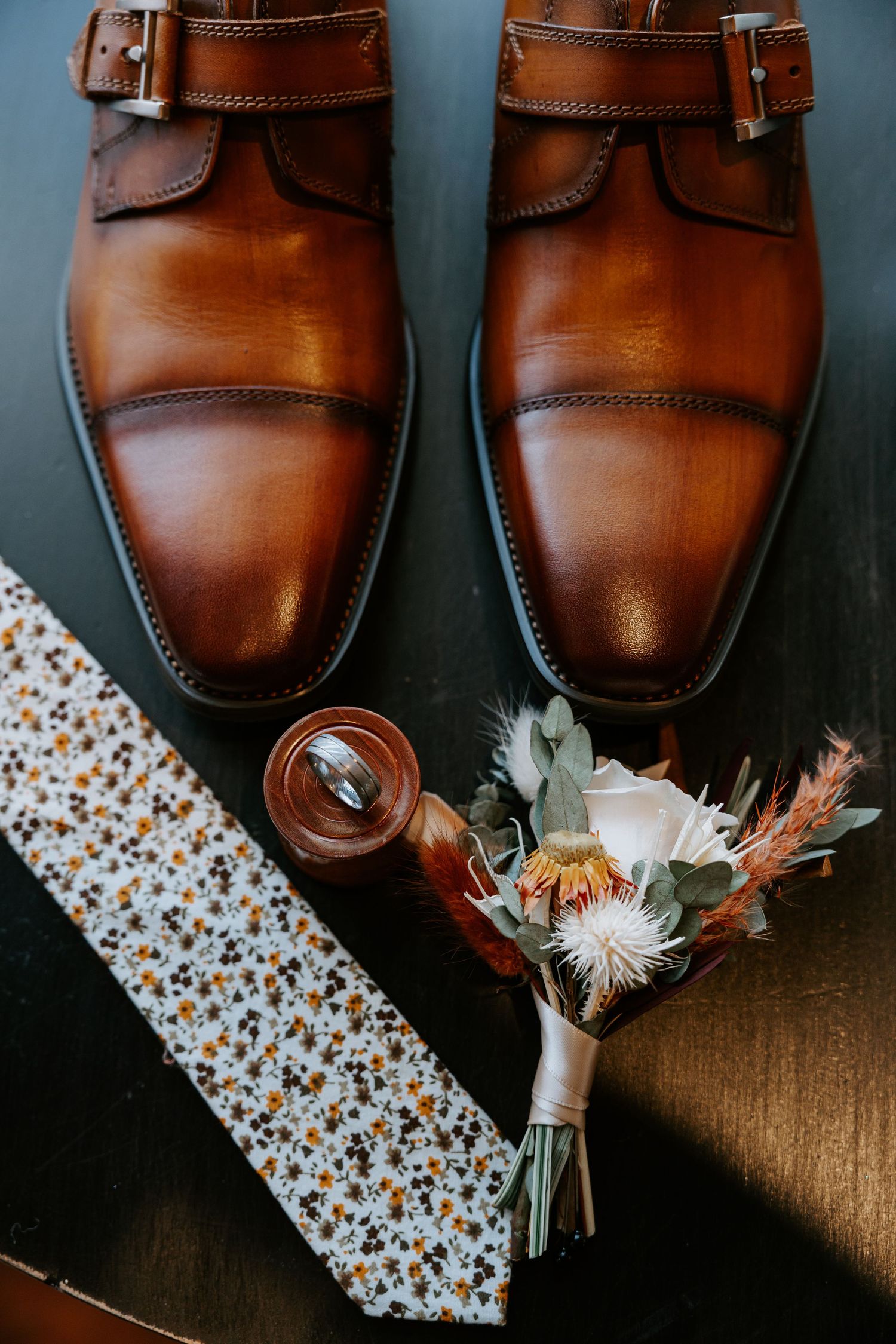 Brown leather dress shoes arranged with floral boutonniere and patterned tie against dark background.