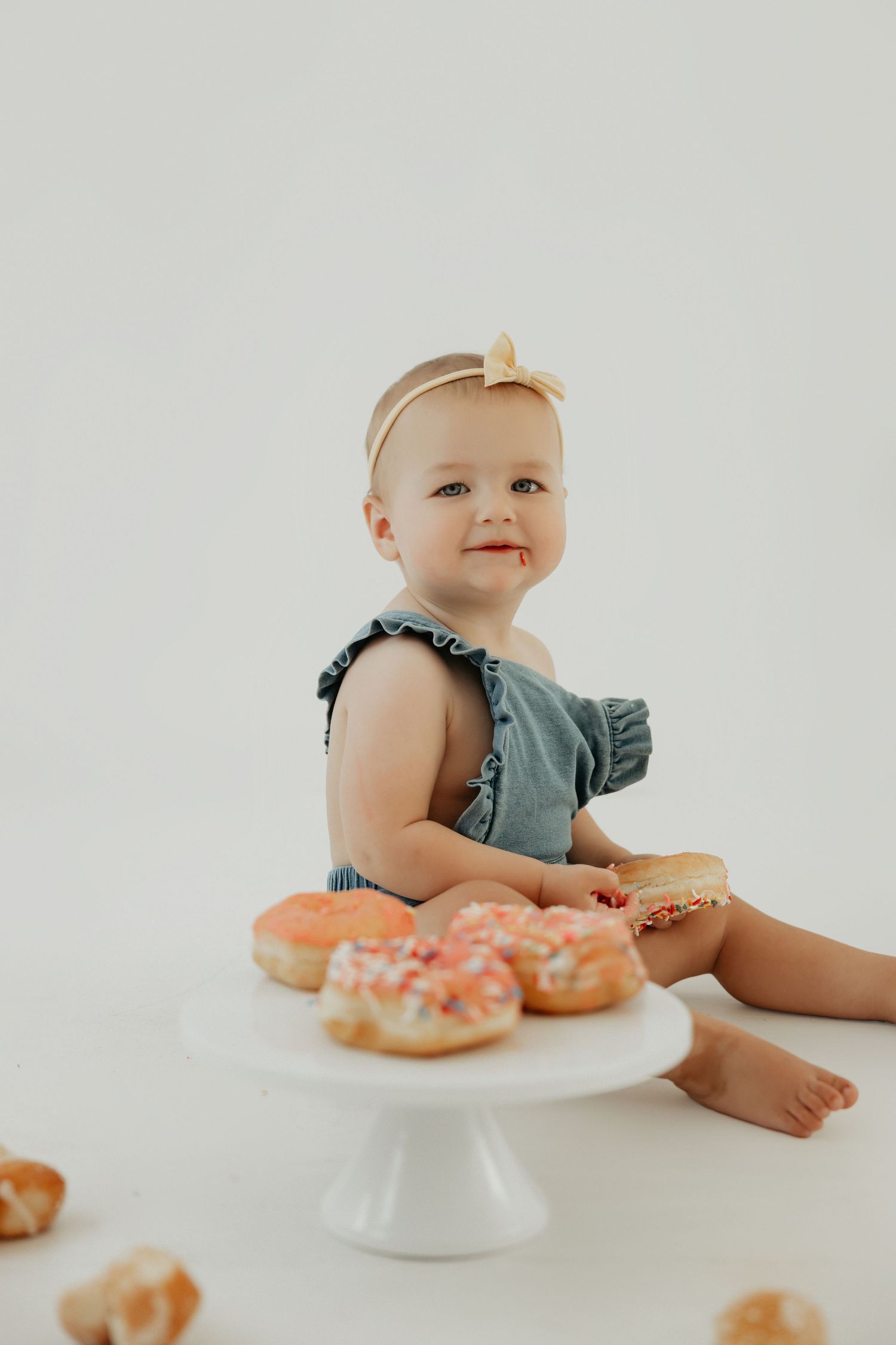 Baby in blue outfit reaches for donuts on white cake stand.