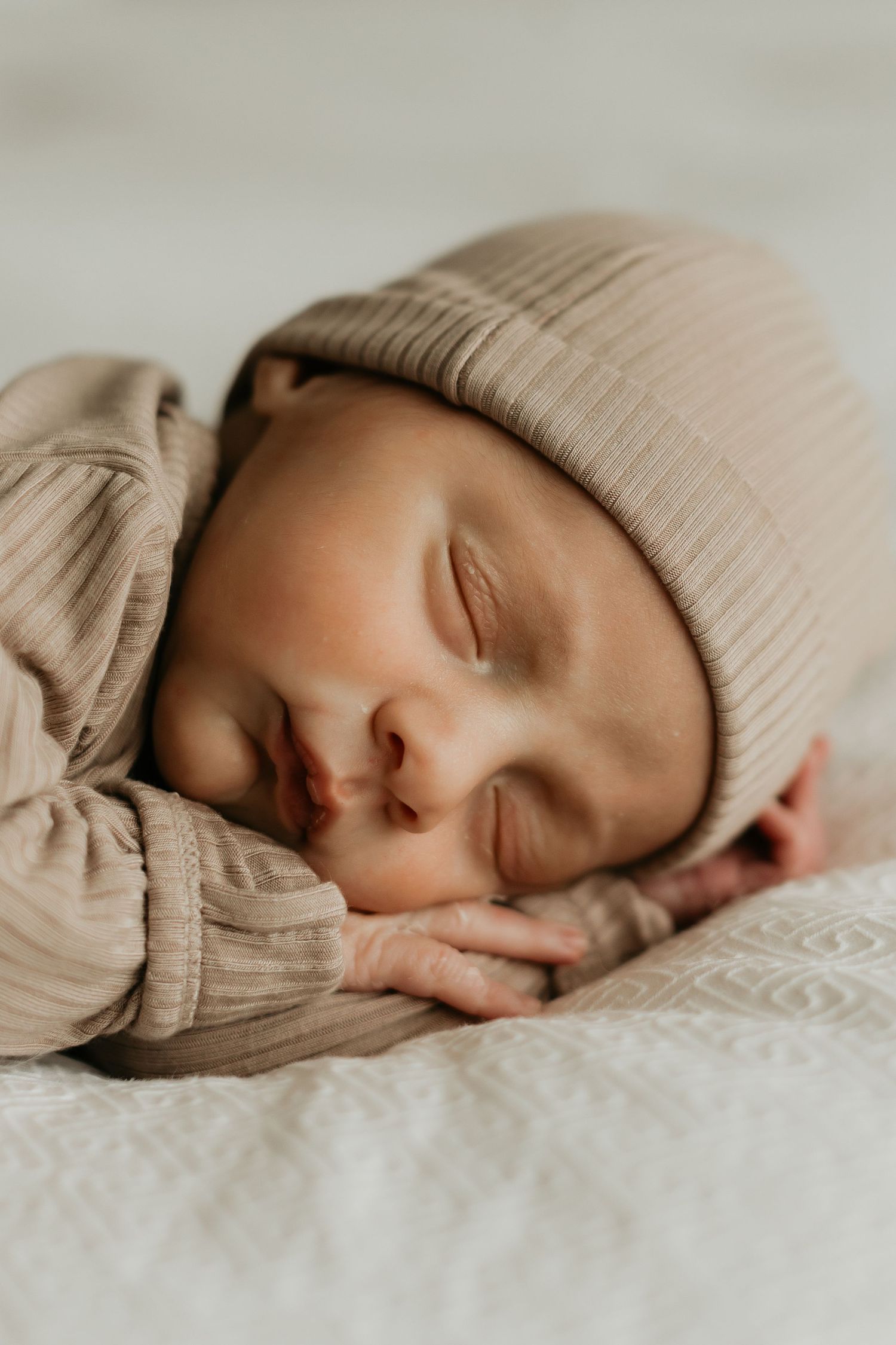 Sleeping newborn in beige knit hat rests peacefully on textured bedding.