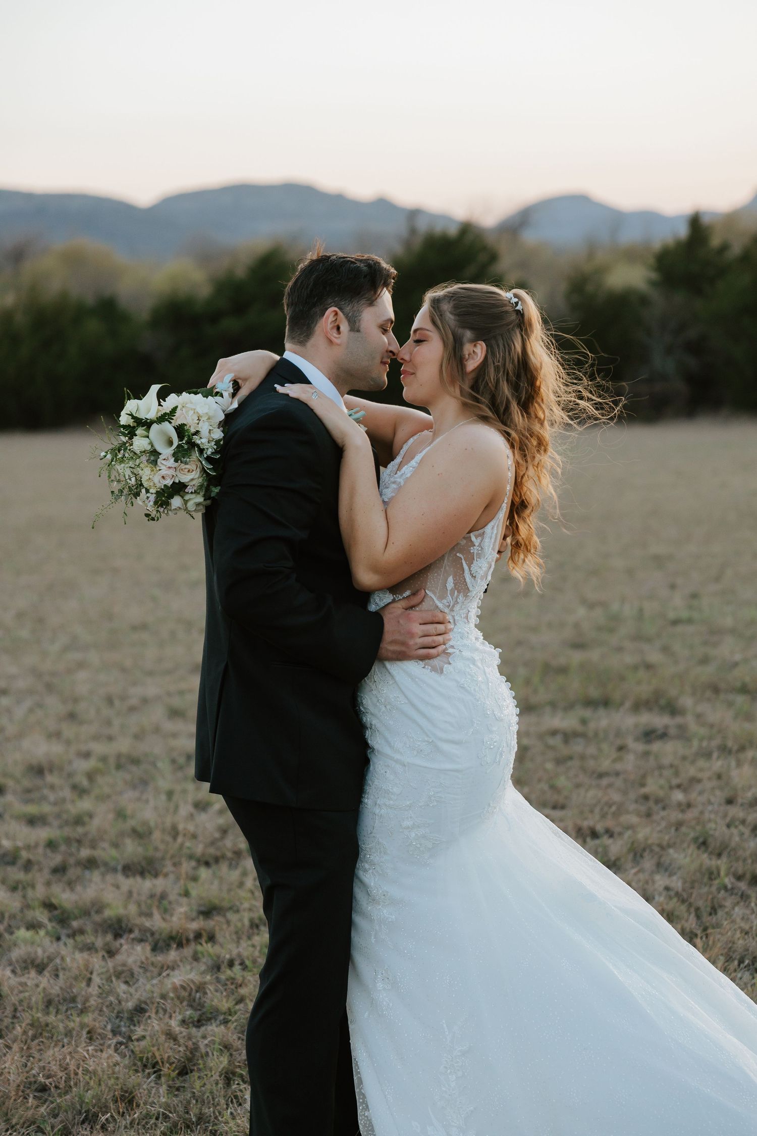 Romantic sunset wedding photos of couple embracing in an open field with mountains in background.