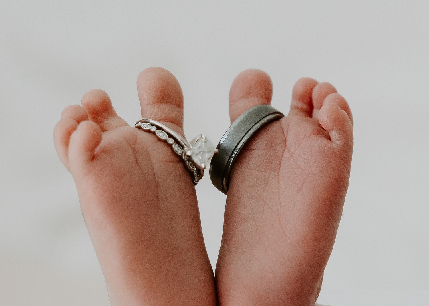 Wedding rings rest between tiny newborn baby feet against a white background.