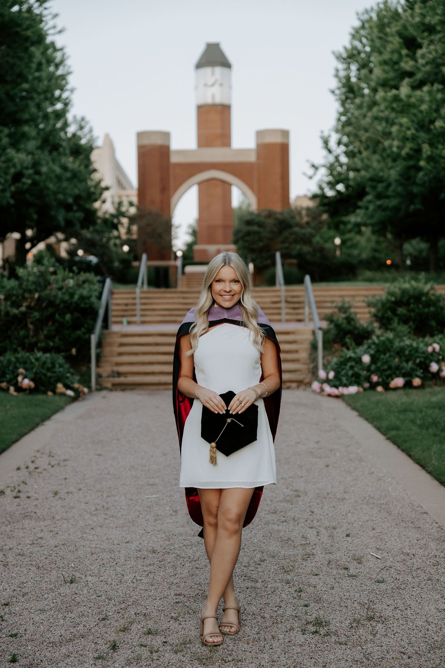 Person in white dress poses playfully on gravel path with brick building in background.