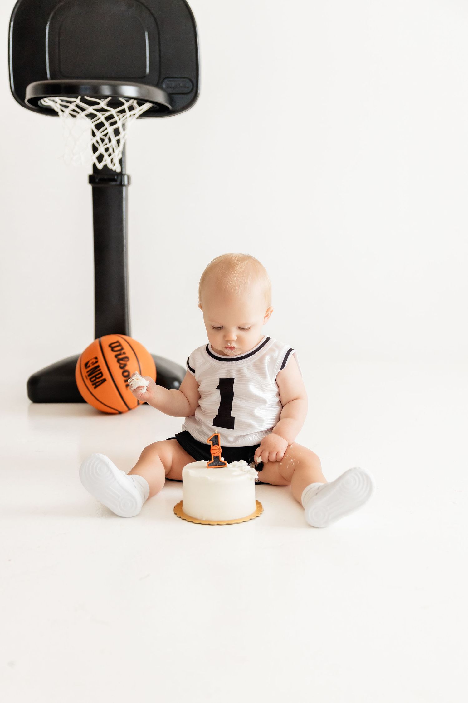 Baby in basketball jersey sits with cake and ball under mini basketball hoop.