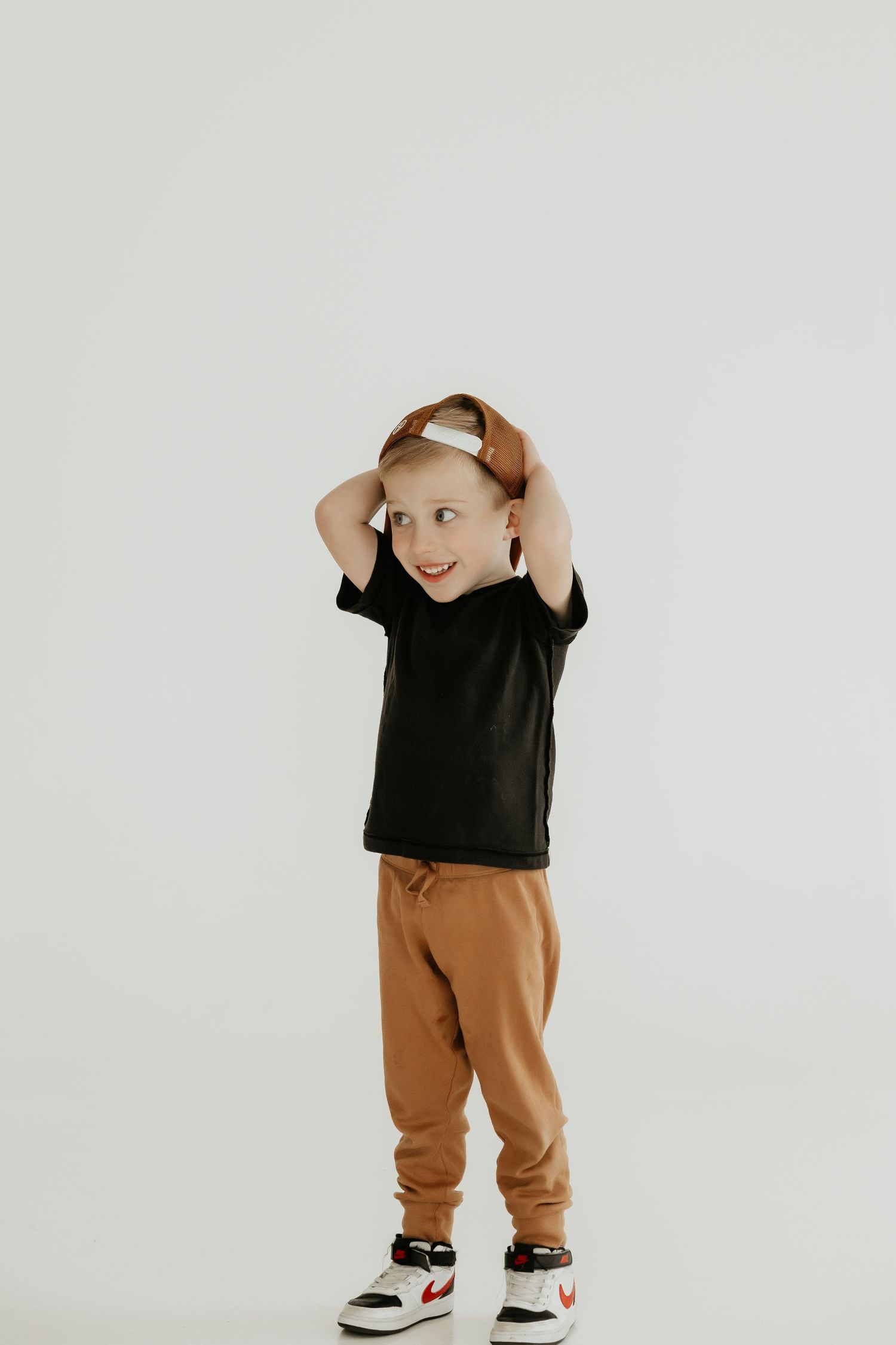 Child in black shirt and brown pants poses playfully against white wall.