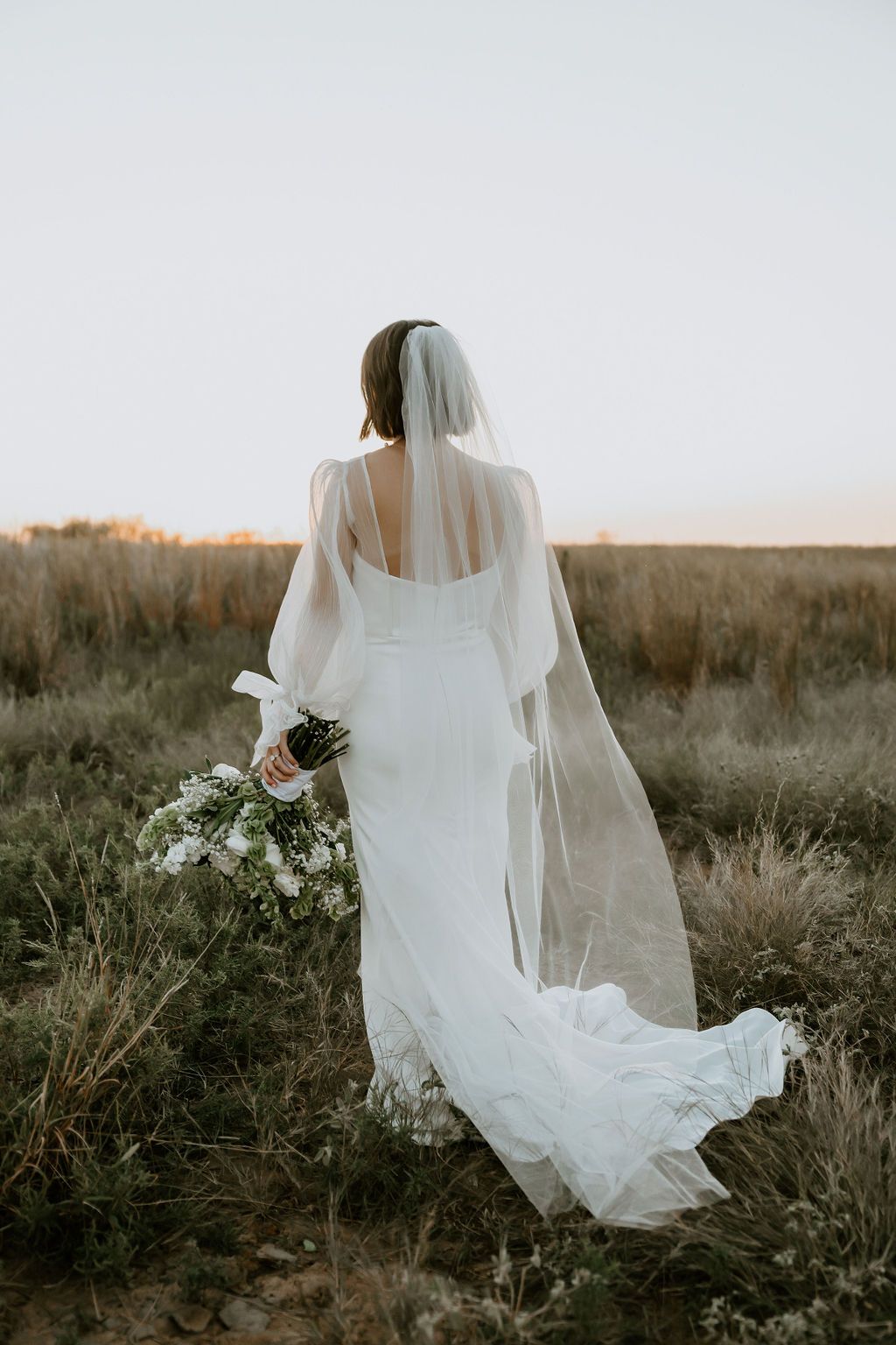 Bride in flowing white dress and veil walking through a rustic field at golden hour.