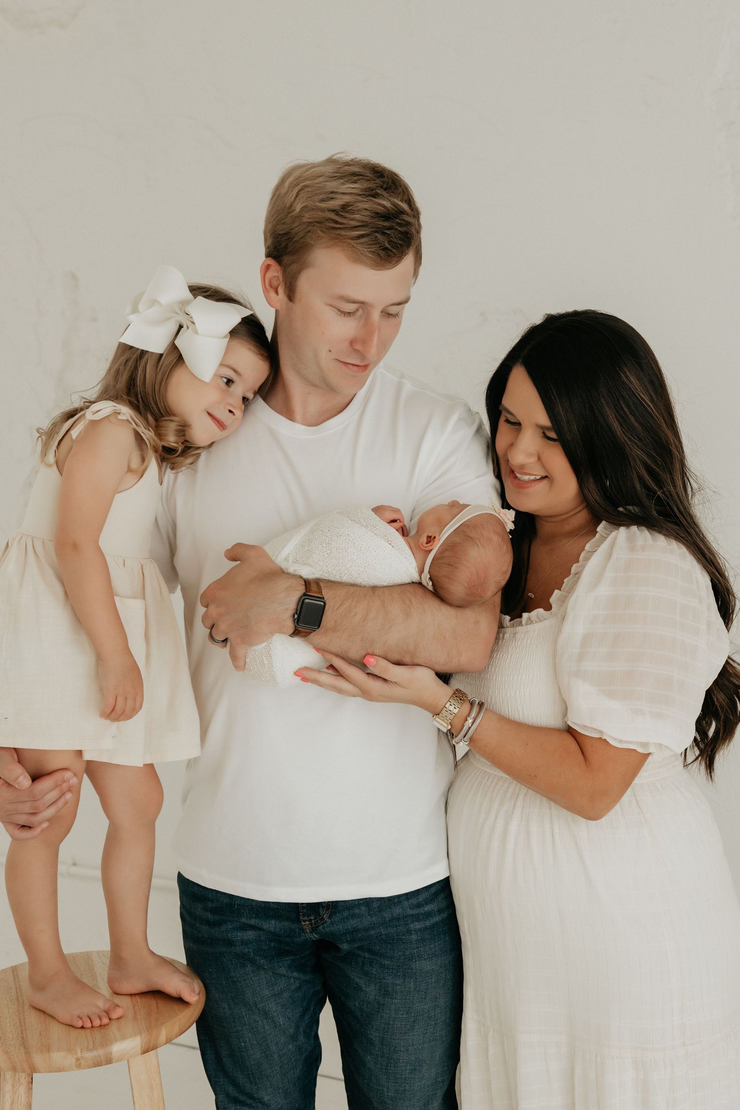 Family in white outfits embracing and holding baby against neutral background.