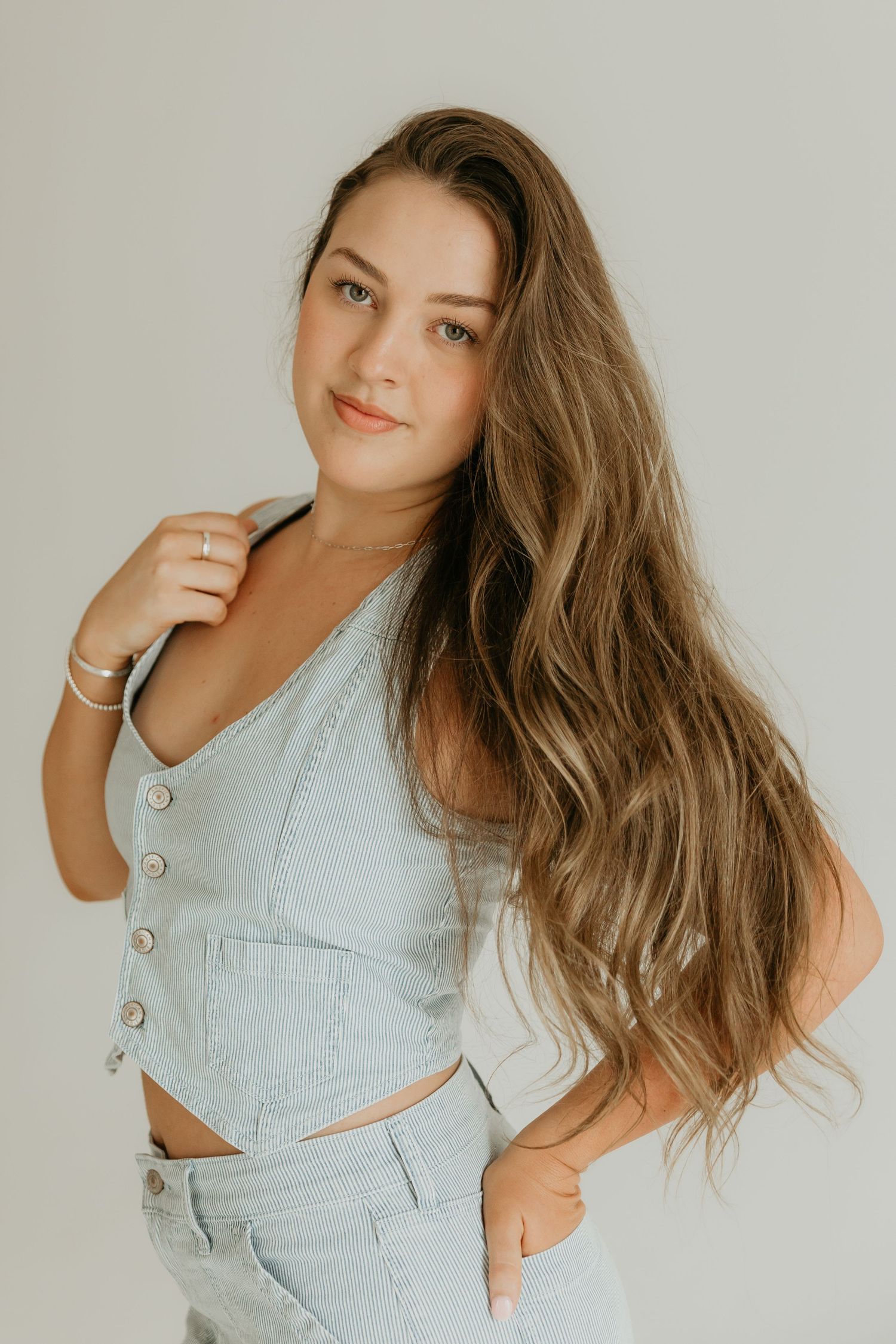 Model in light denim outfit poses against white wall showcasing long wavy hair.