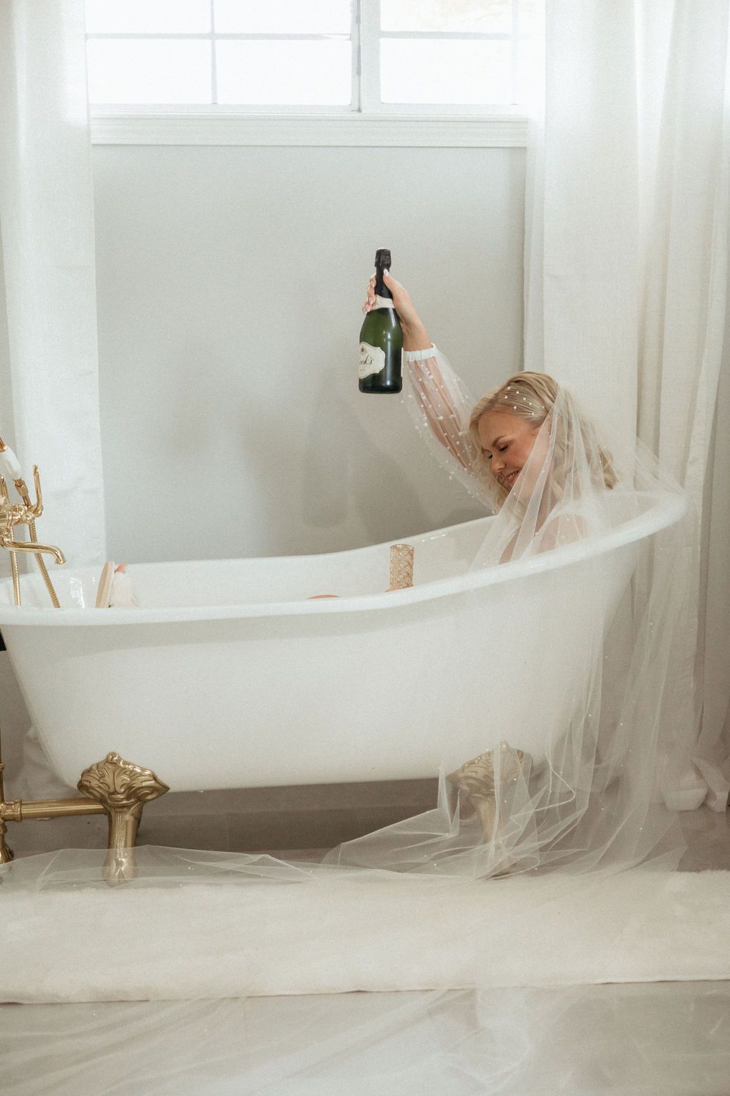 Artistic bridal portrait in white bathtub with flowing veil in bright studio setting.
