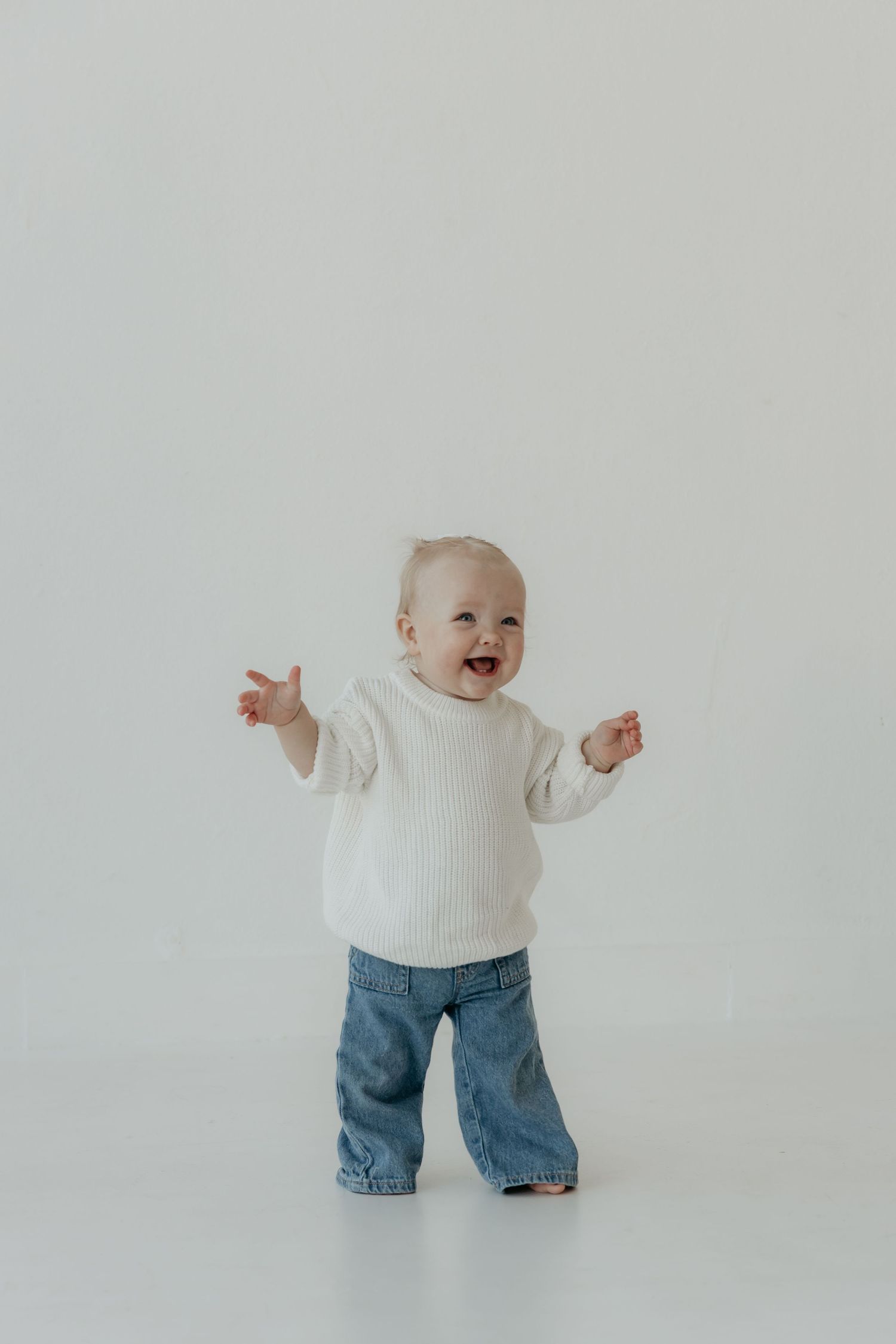 Baby in white sweater and jeans stands against white background with arms raised.