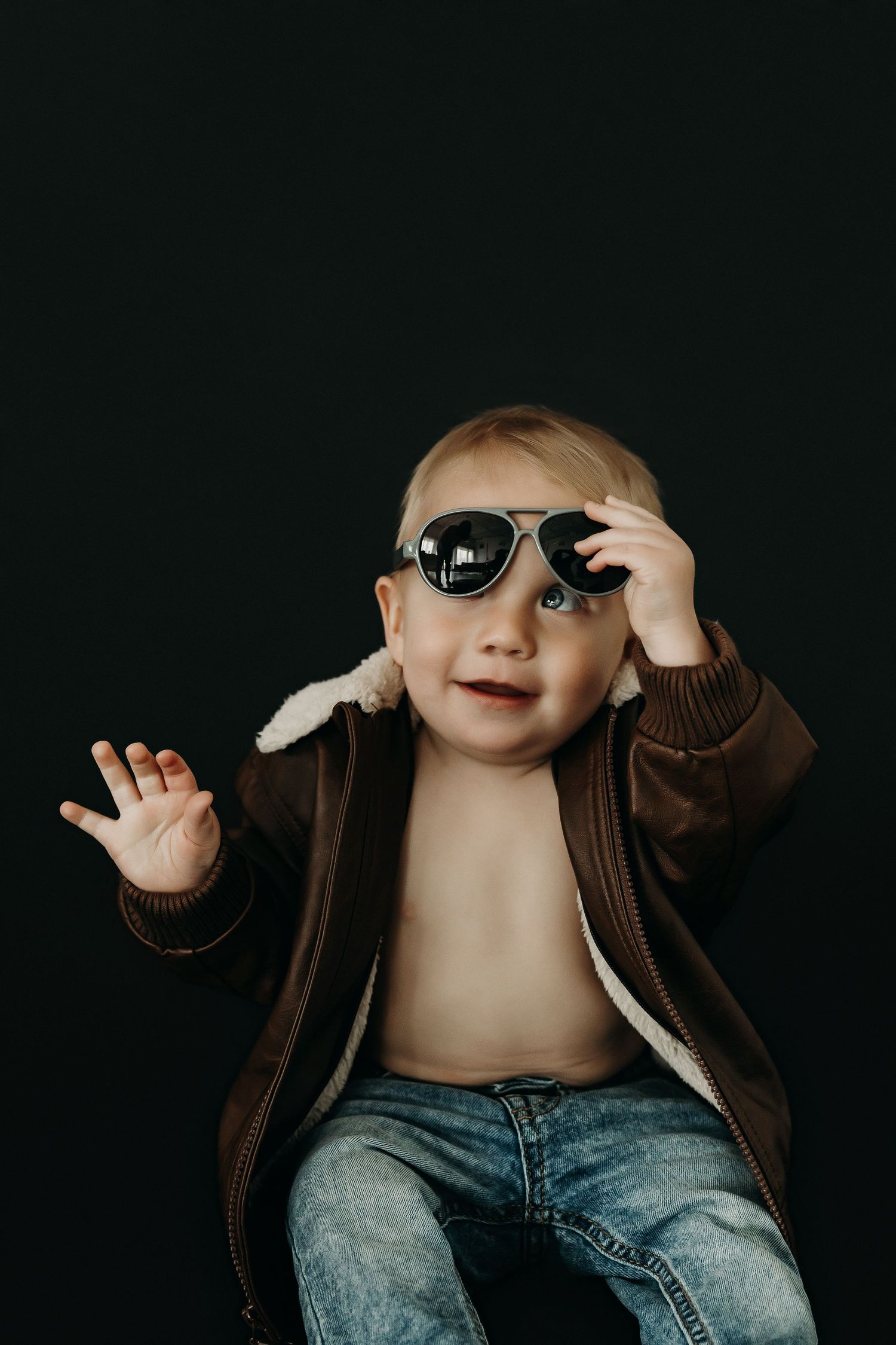 A baby wearing sunglasses and a brown jacket poses playfully against a dark background.