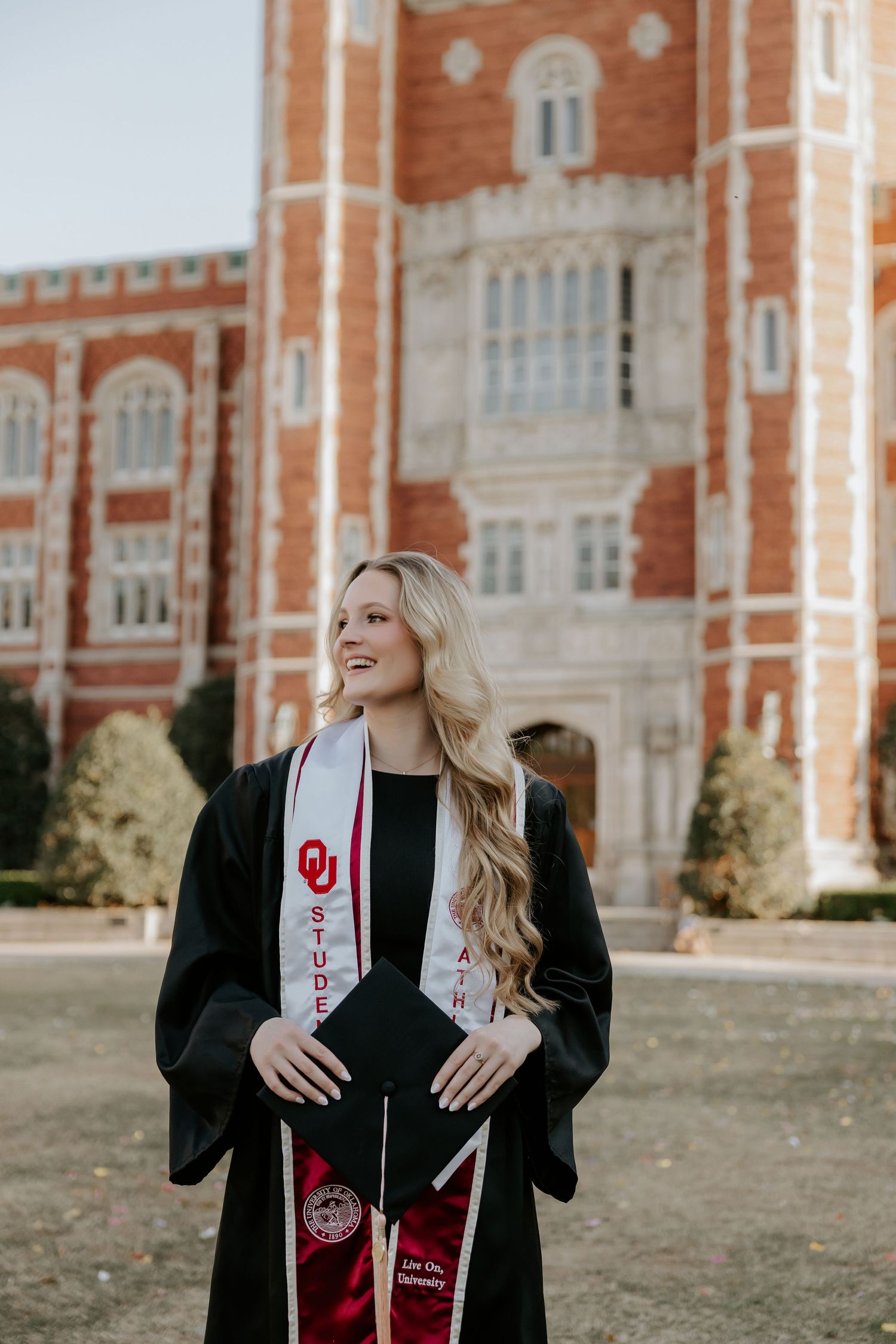 Graduate in black gown with red stole posing in front of brick university building.