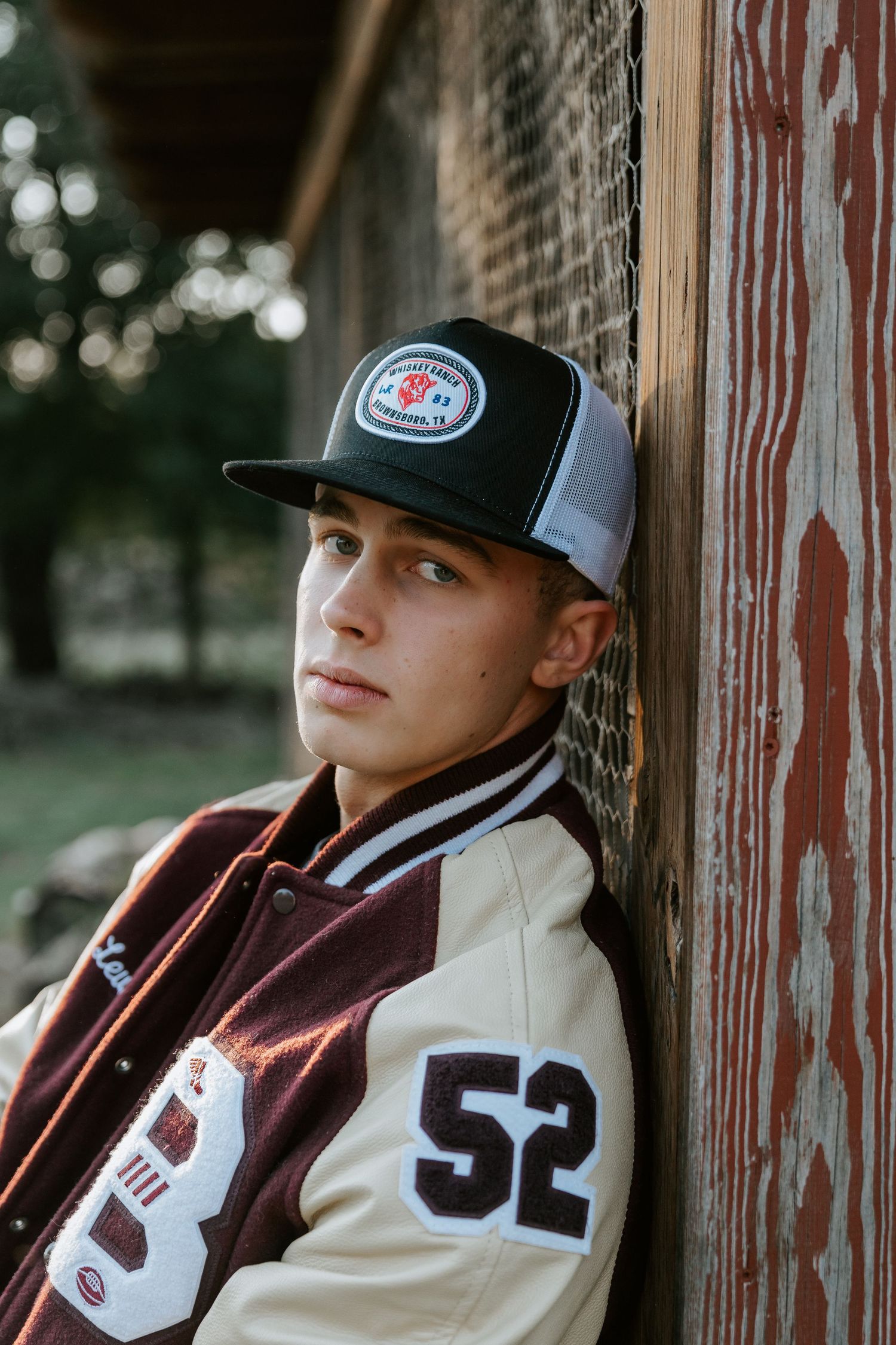 Sports fan wearing varsity jacket and baseball cap poses against rustic wooden backdrop.