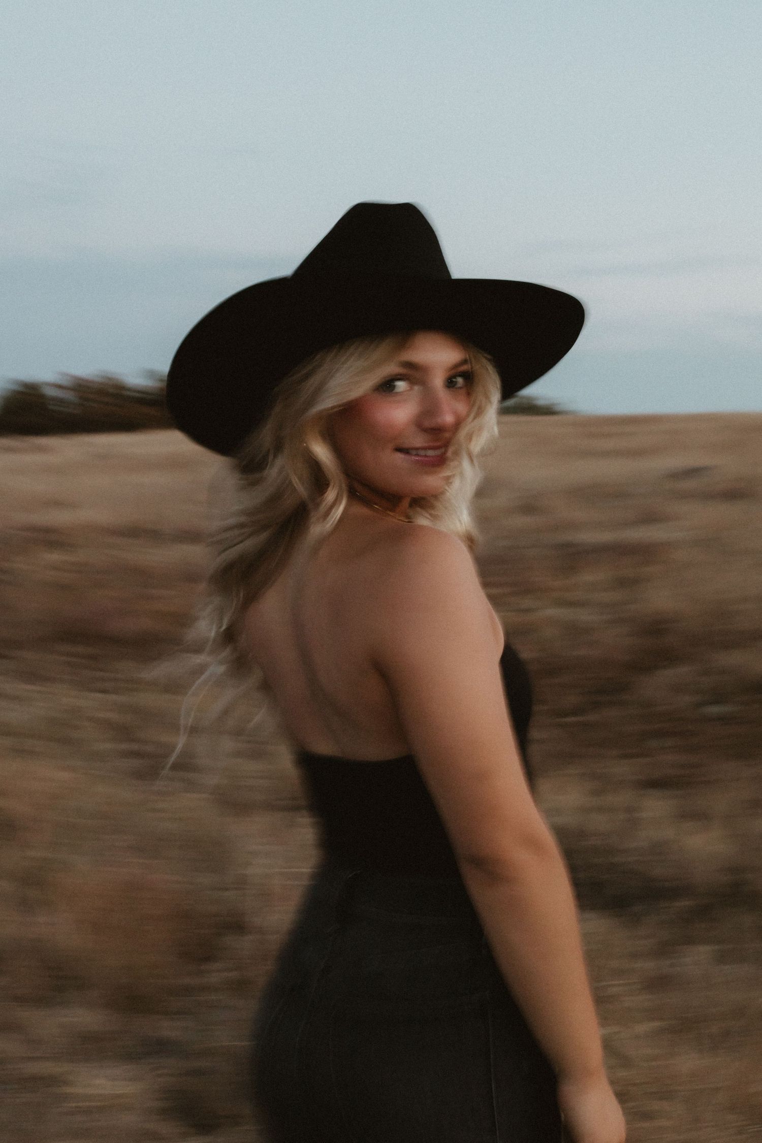 Person wearing black hat and dress poses in desert setting.