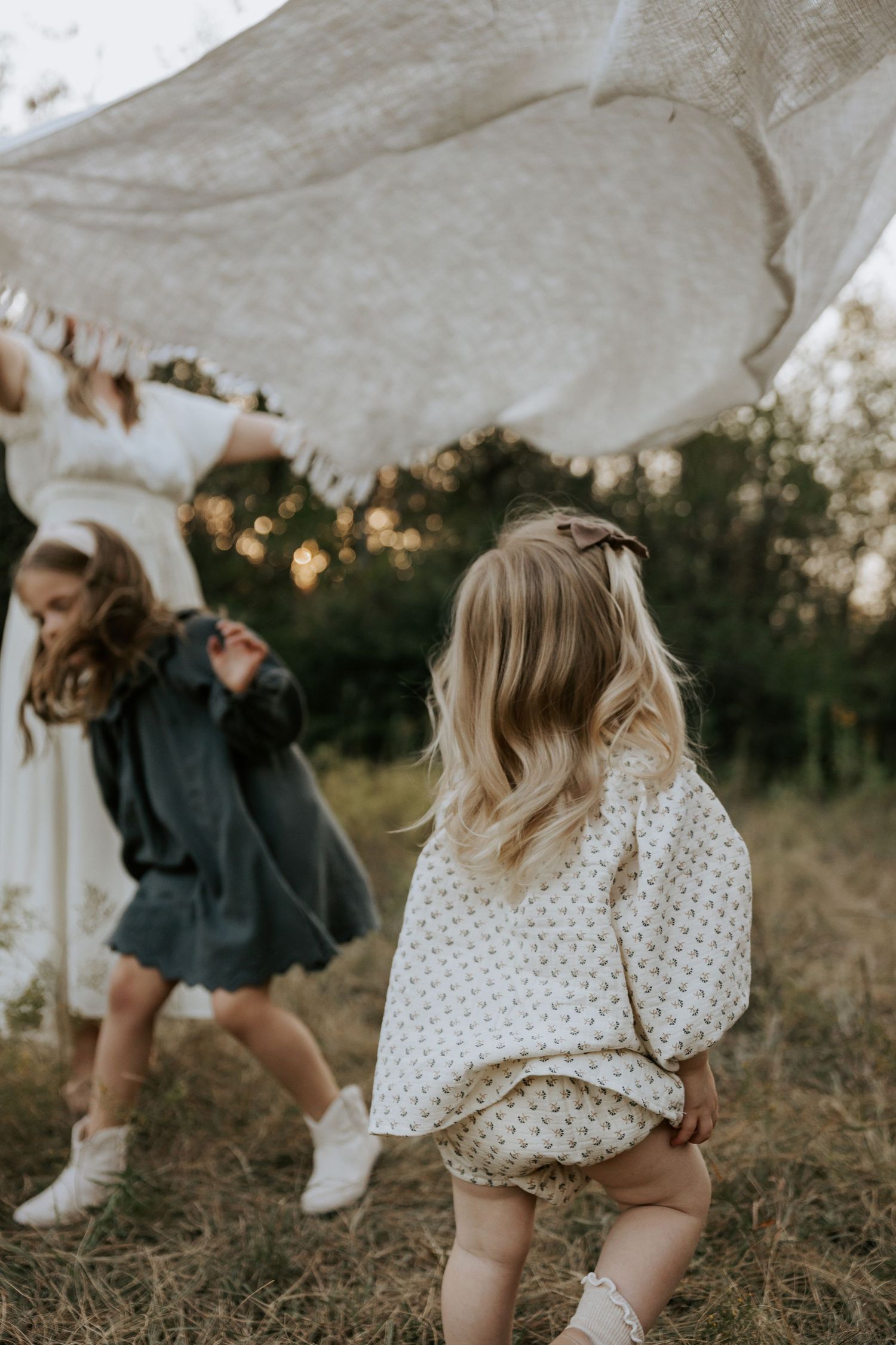 Child plays under flowing white fabric in rustic outdoor setting at sunset.