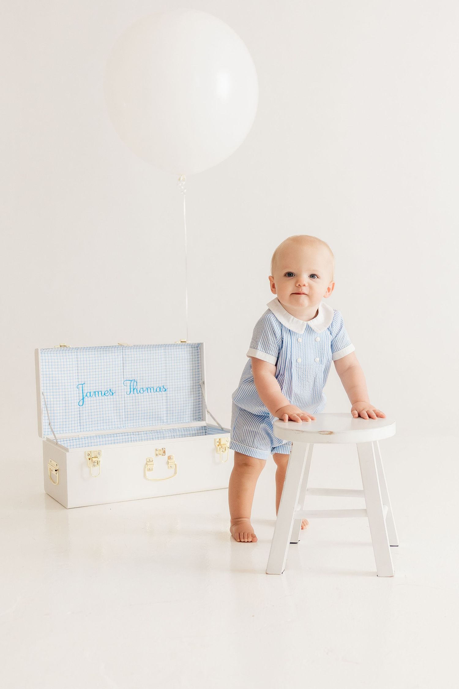 Baby in blue romper stands by white stool with decorative box and balloon in background.