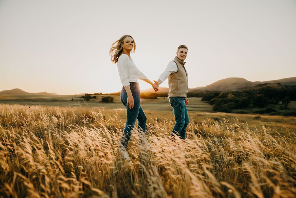 A couple walks hand in hand through tall golden grass at sunset.