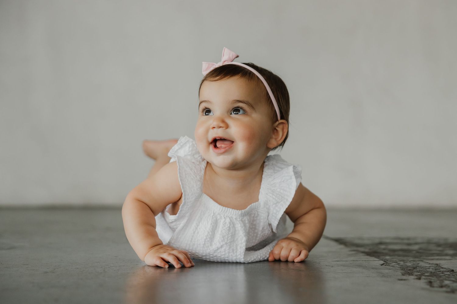 Baby in white dress lying on gray background during photoshoot.