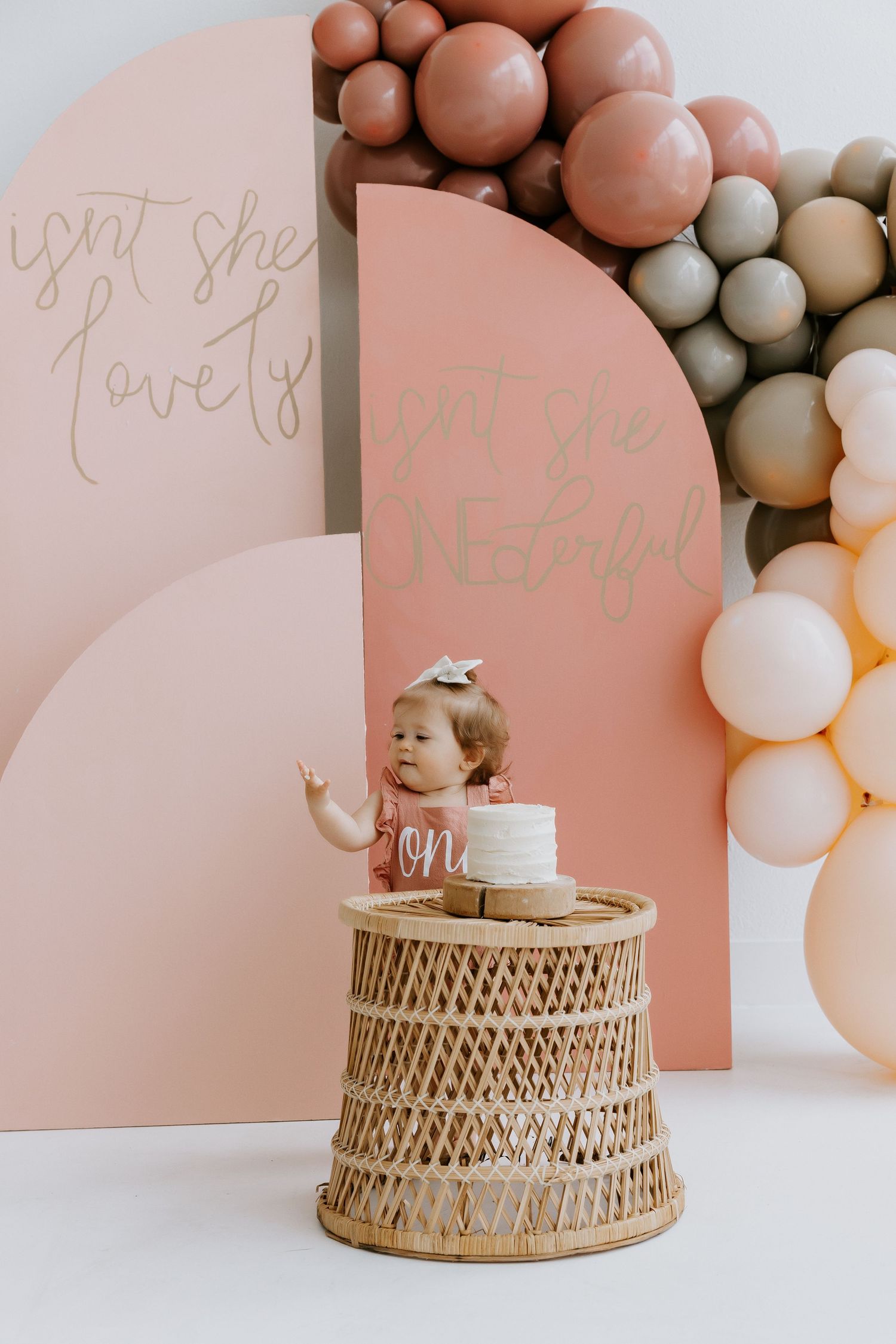 First birthday setup with pink backdrop, balloon arch, and woven pedestal.