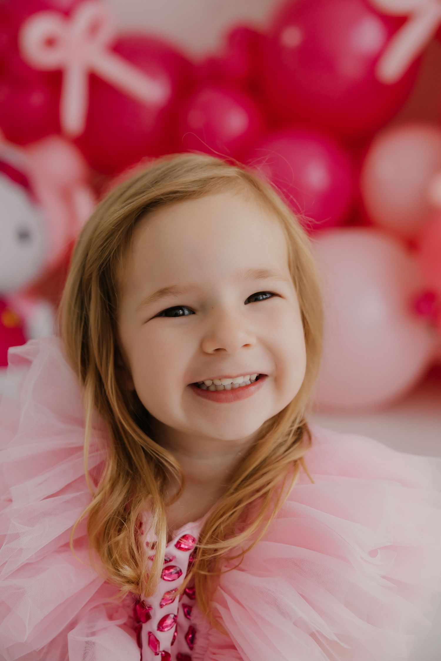 Smiling child wearing pink outfit against pink balloon backdrop.