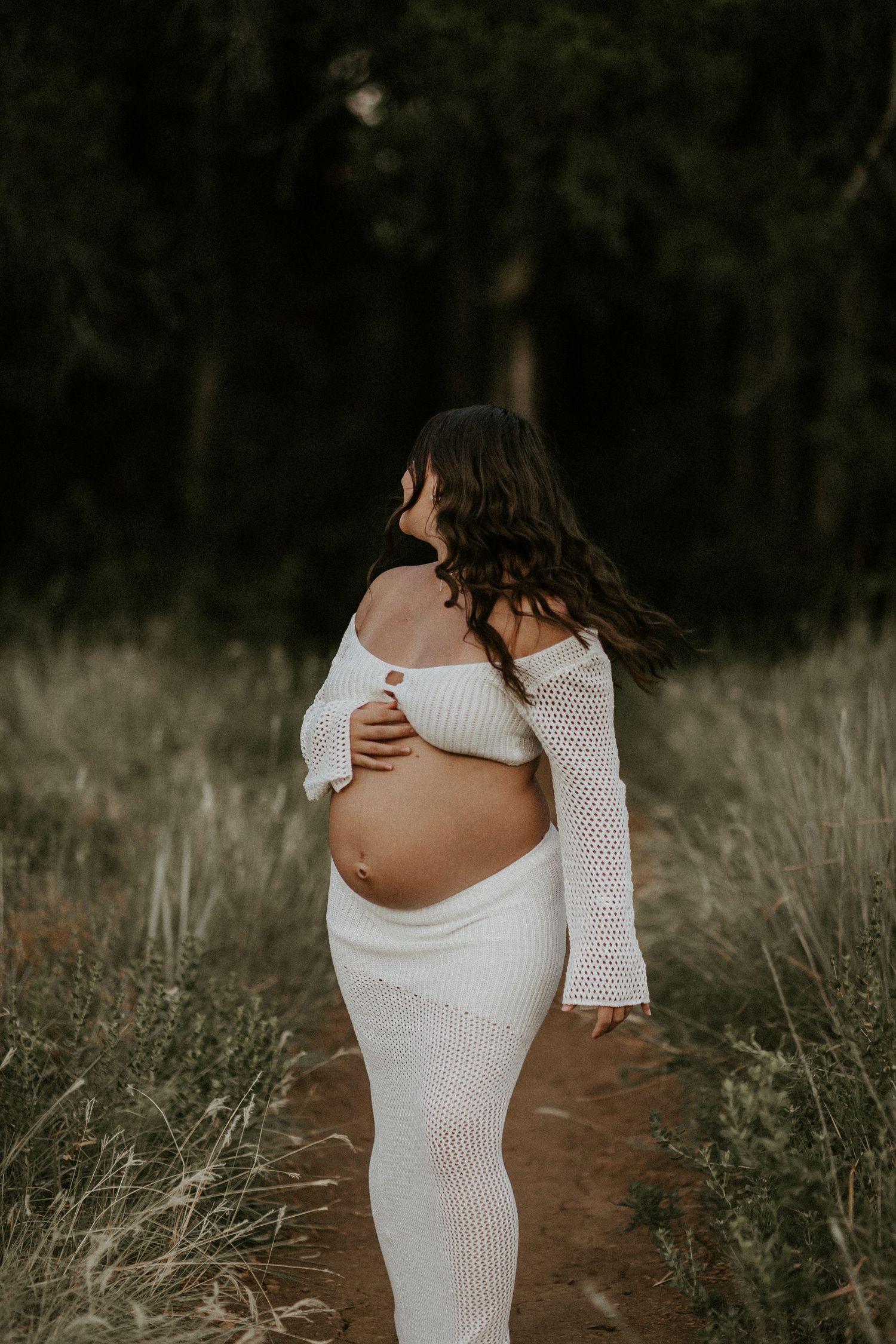 Dramatic maternity photos in a white knit dress on a forest path at dusk.