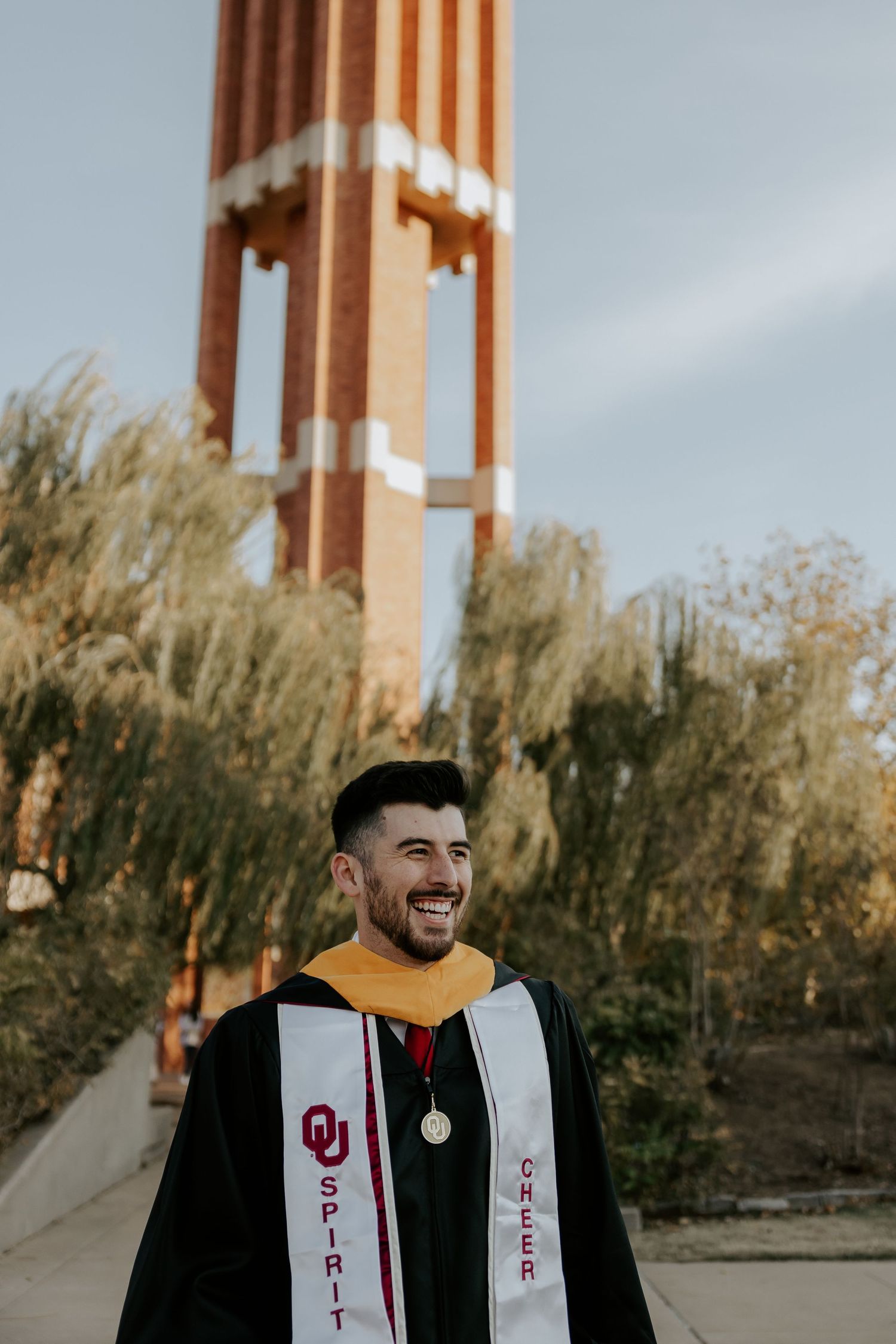 Graduate in academic regalia poses outdoors with architectural columns in background.