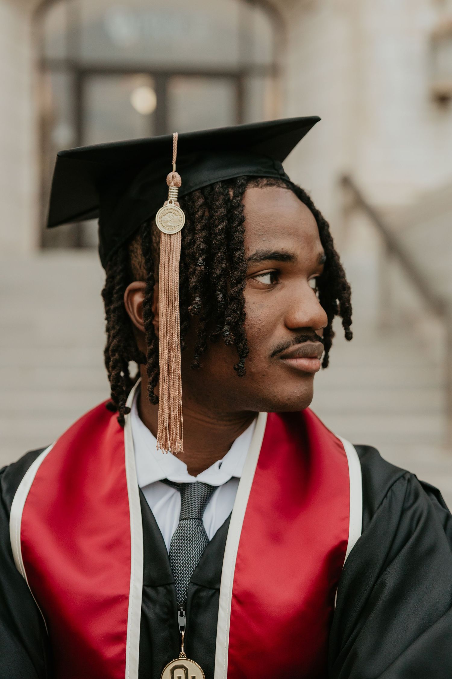 Graduate wearing black cap with gold tassel and red stole against neutral background.