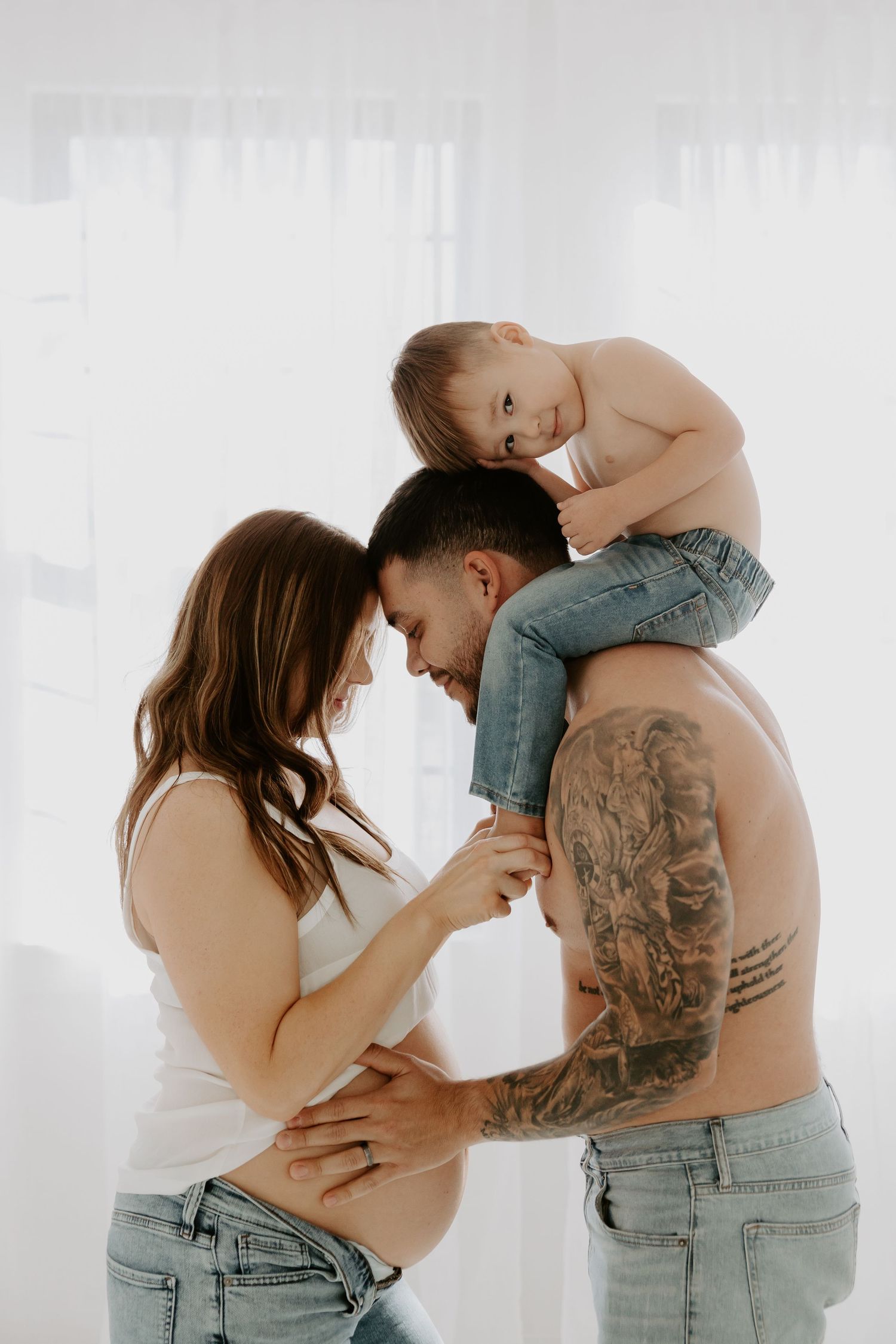 Intimate maternity photoshoot featuring couple in casual denim against white backdrop.