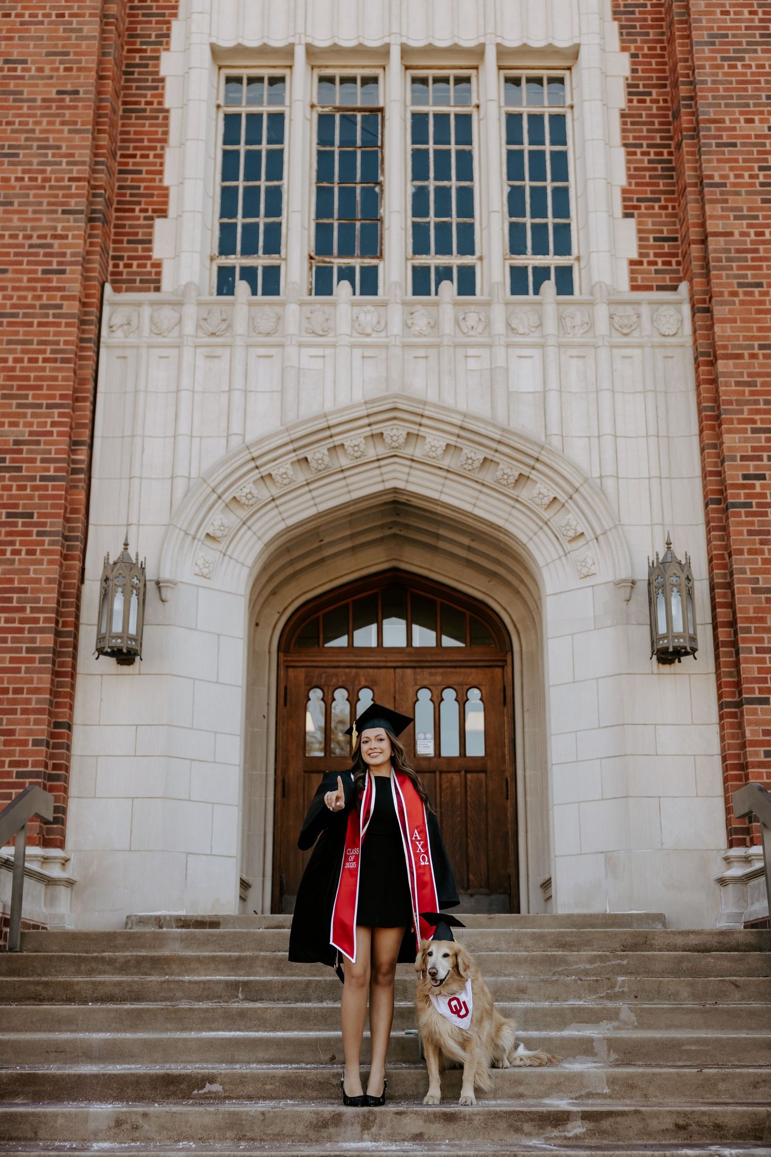 Graduate and their Golden Retriever sit on stone steps in front of a brick university building entrance.