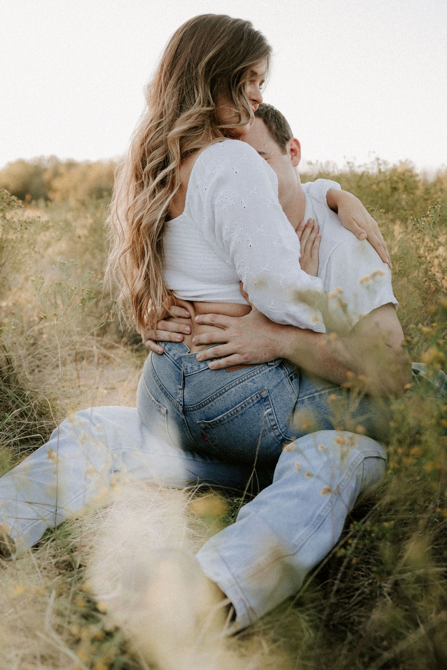 Couple in casual denim embracing in sunlit field with wild grasses.
