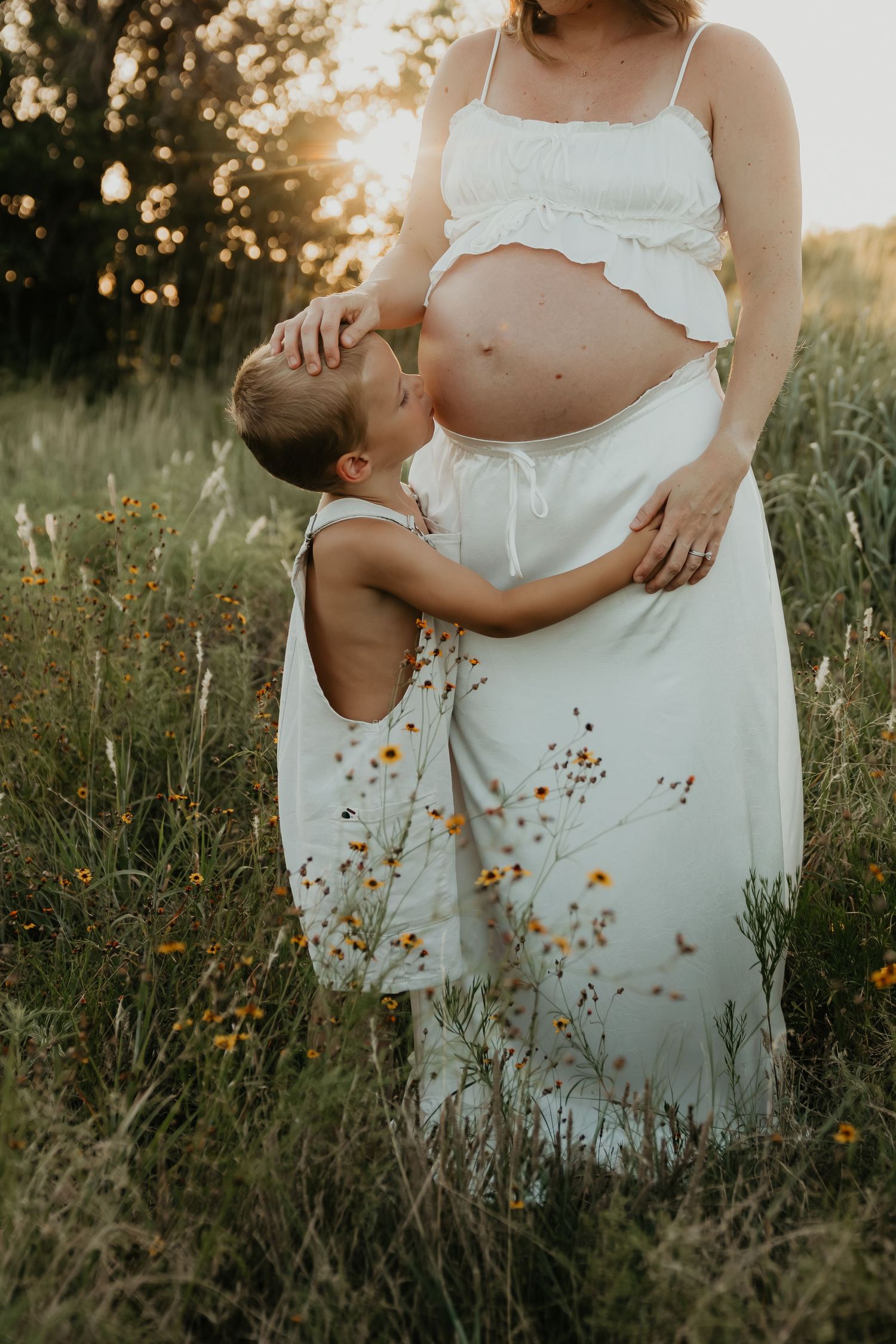 Tender maternity photo of pregnant woman in white dress with child embracing her belly at sunset.