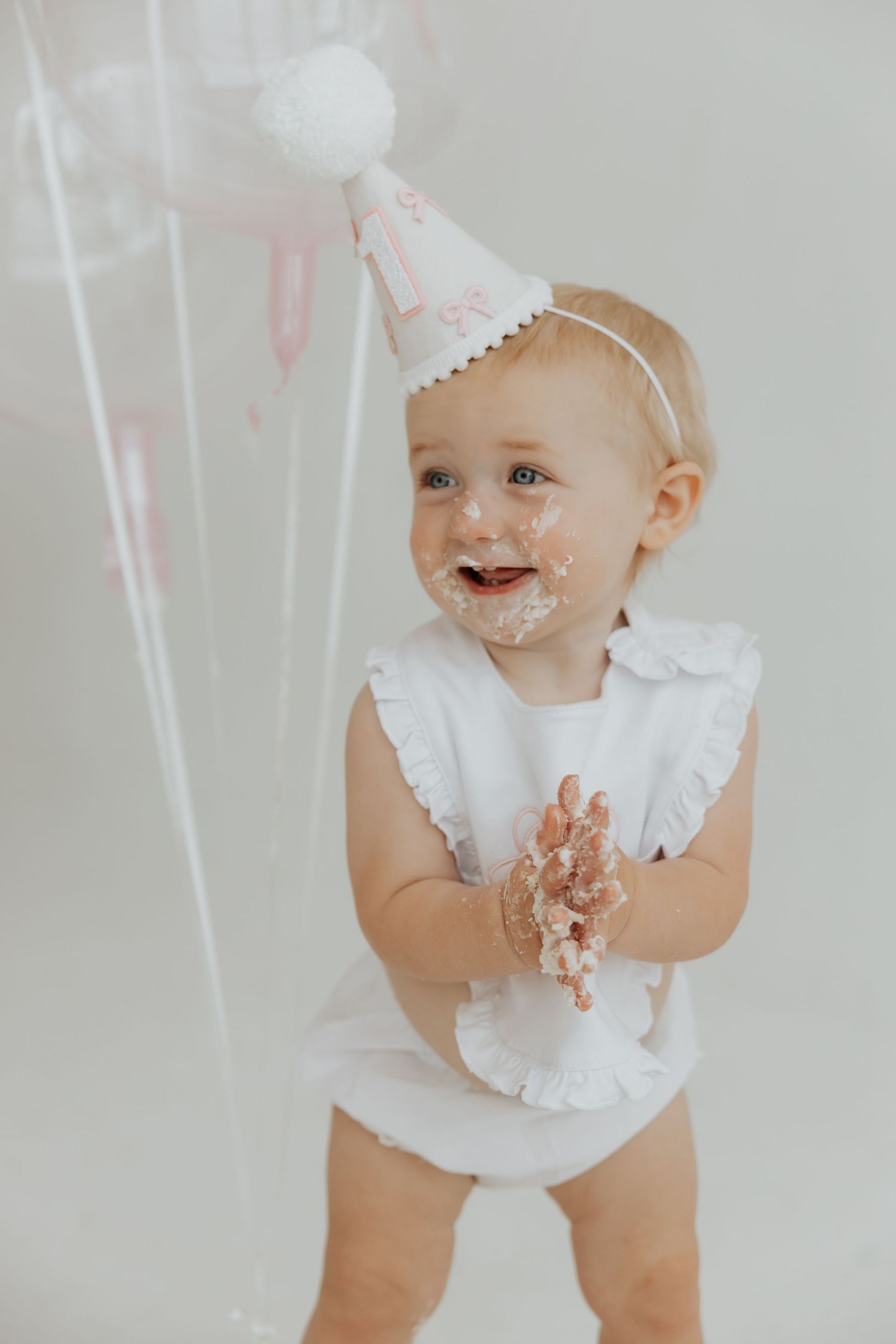 Baby in white outfit gets messy with cake frosting during photoshoot.