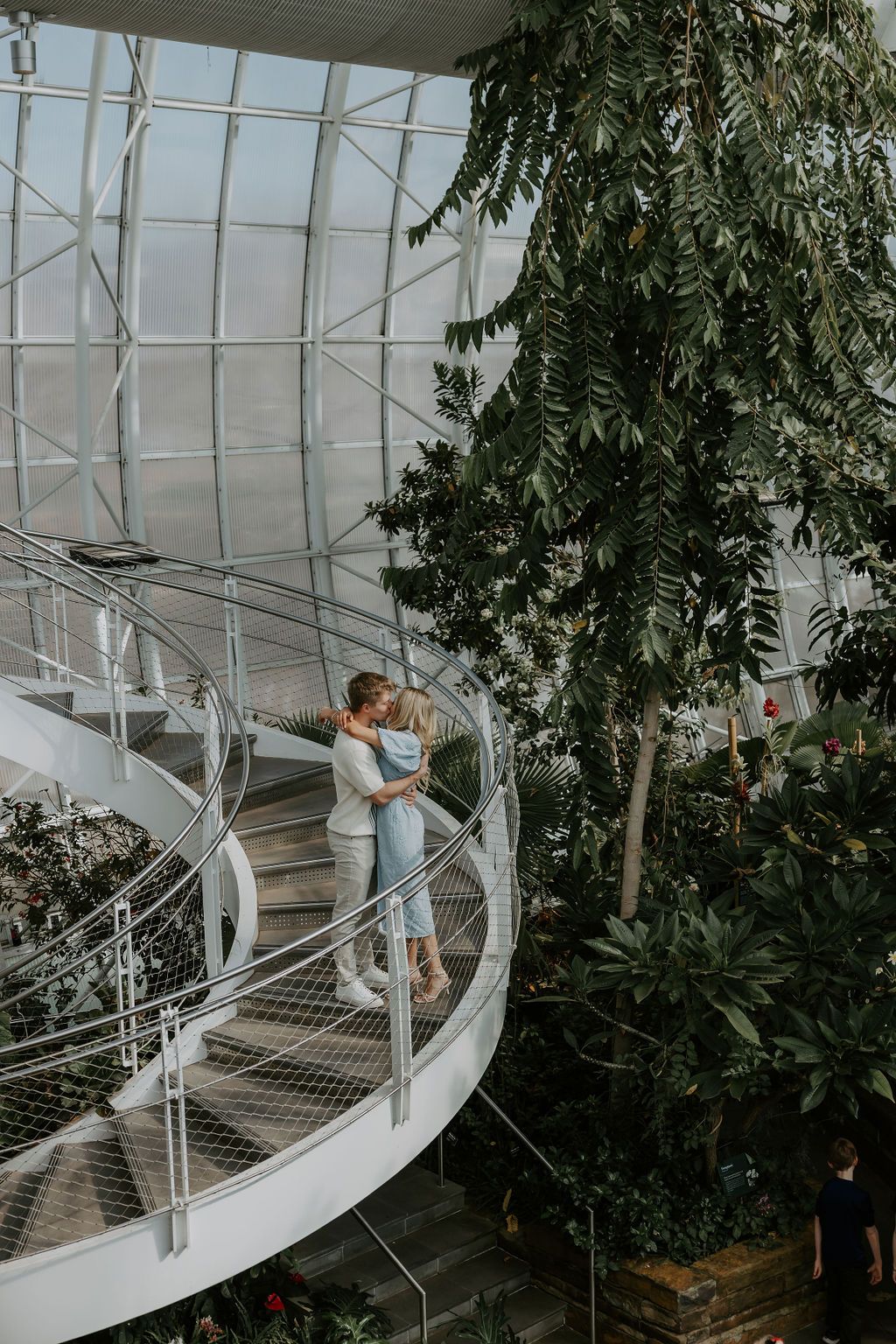Couple posing playfully on curved white staircase in greenhouse conservatory.