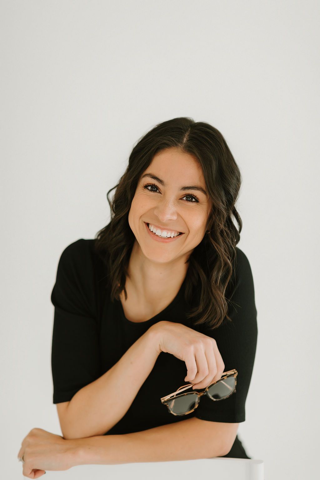Professional headshot of someone holding sunglasses and smiling against white background.