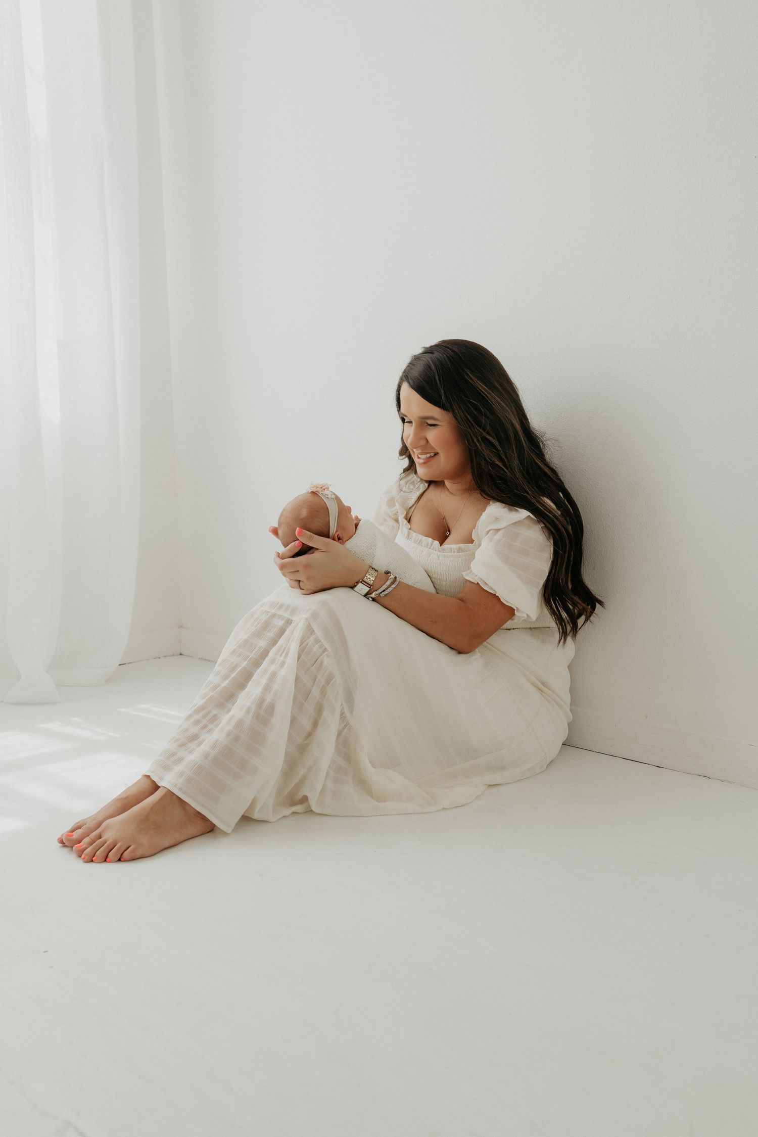 Mother in white dress lying on floor bonding with baby in minimalist setting.
