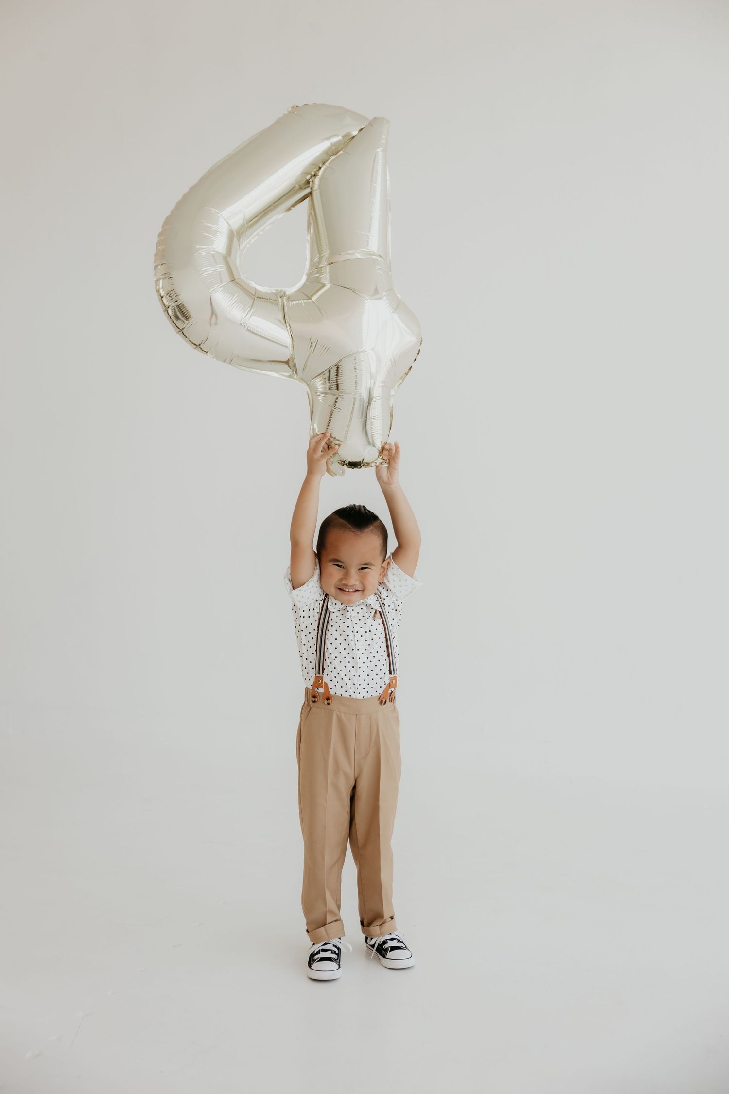 Small child in tan overalls holds a silver balloon number against white background.