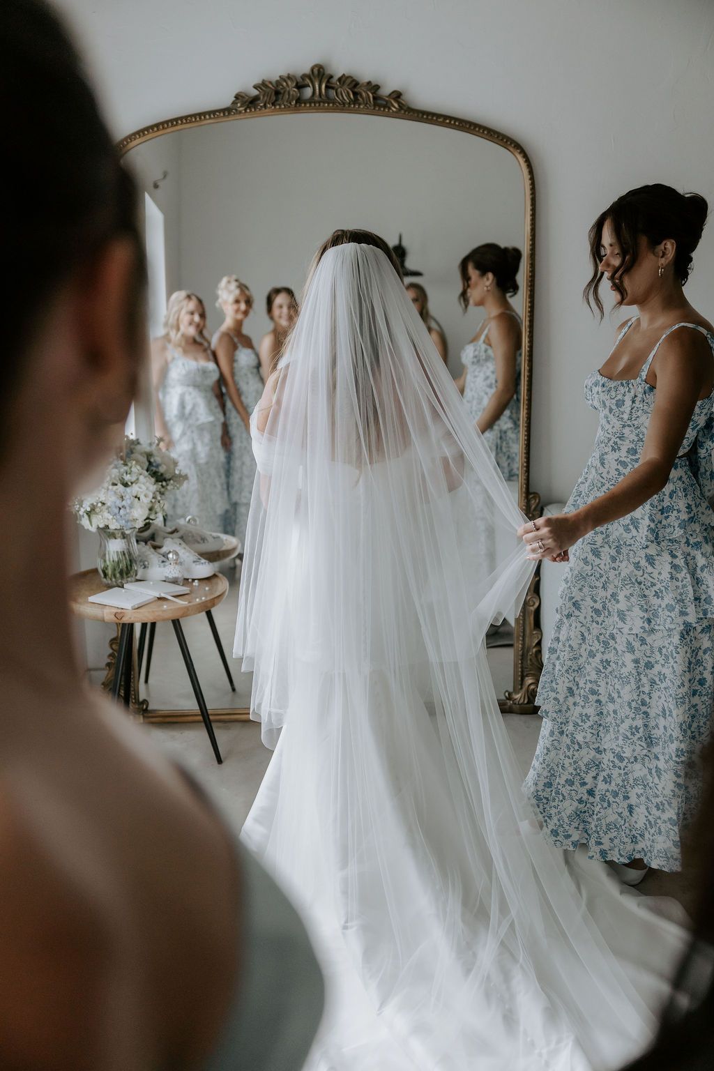 Bridesmaids in floral dresses help adjust a bride's flowing white wedding gown in front of an ornate mirror.