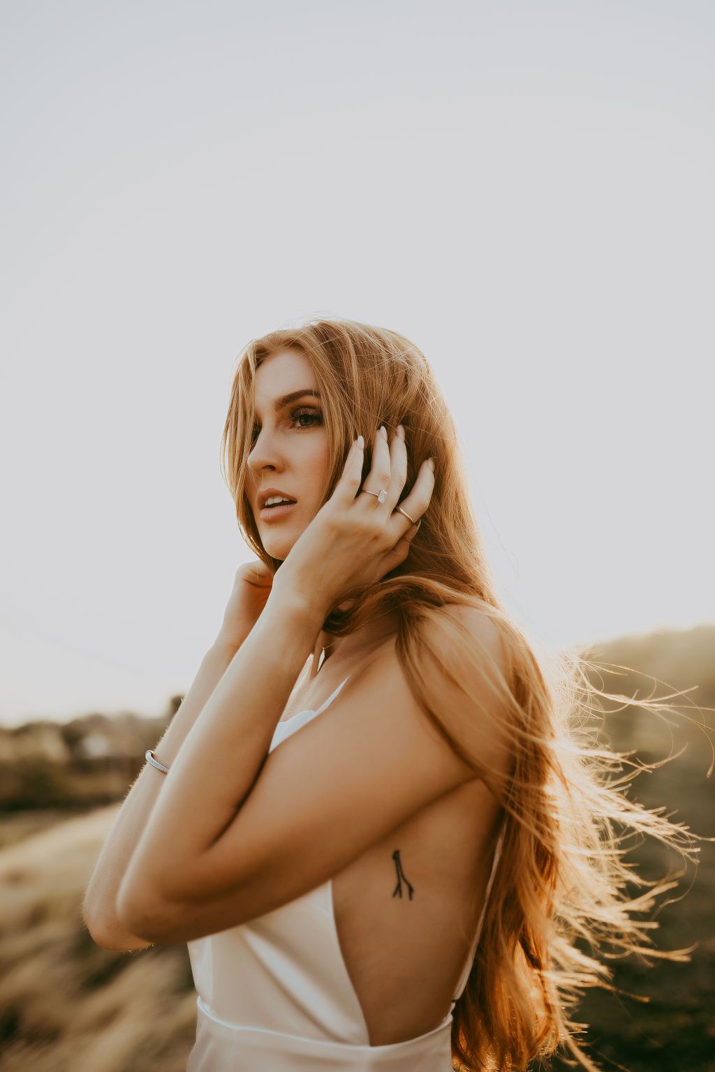 A woman in white poses joyfully at sunset with windswept hair.