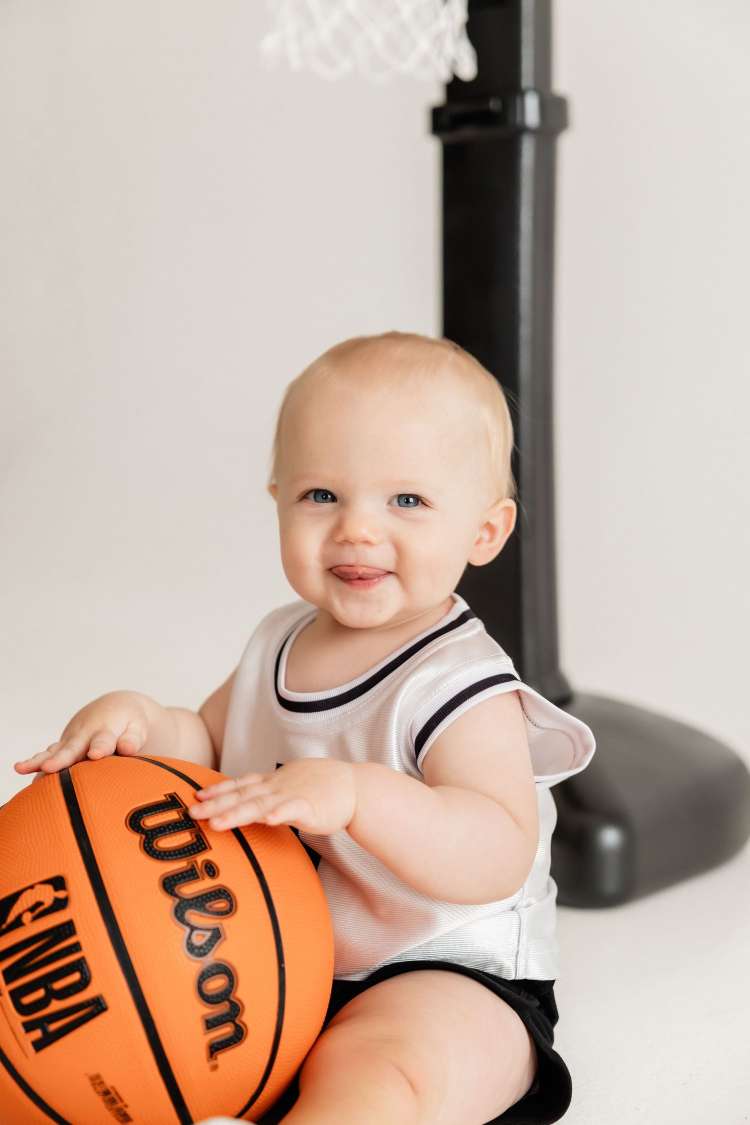 Smiling baby in white jersey holds orange basketball near portable basketball hoop.